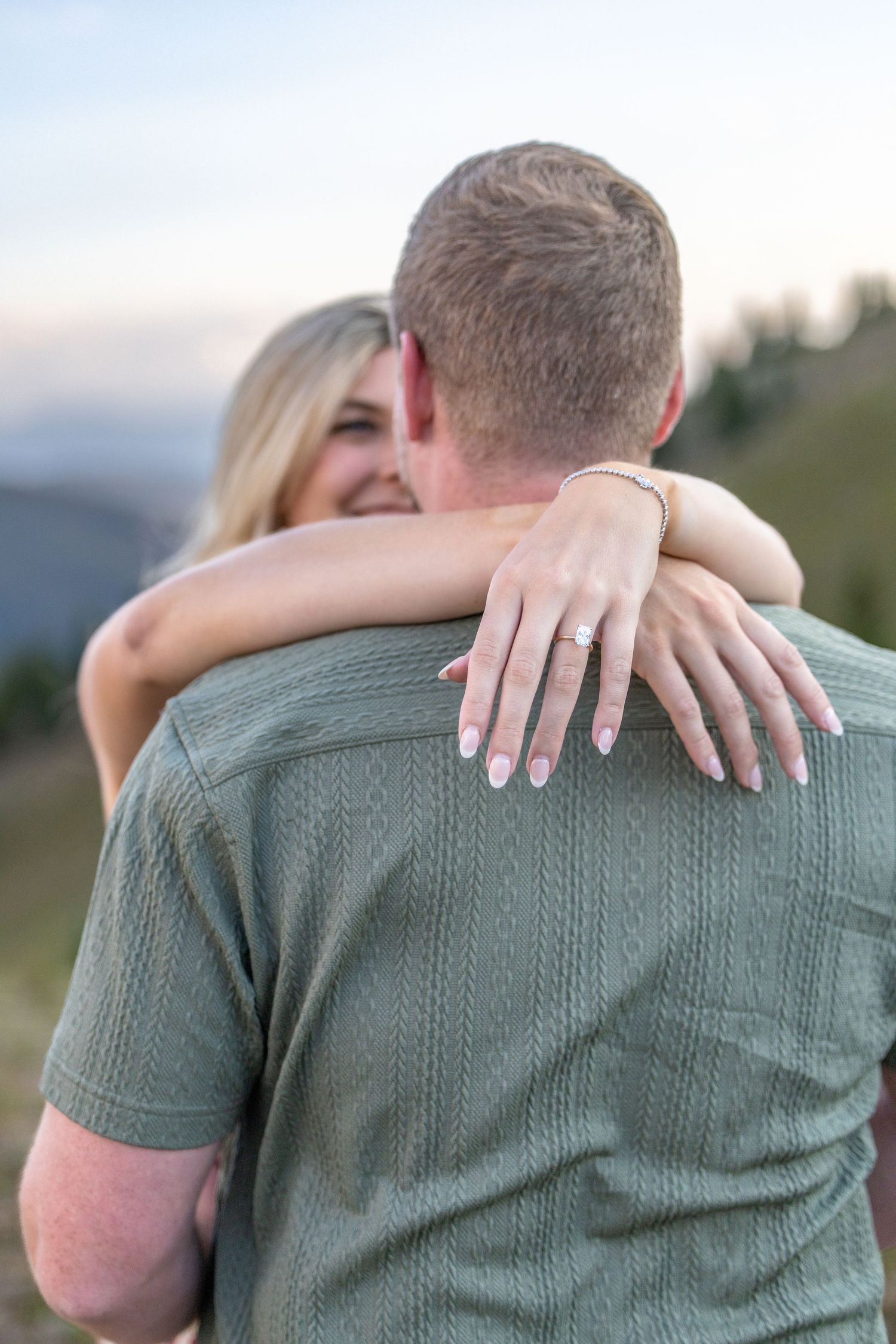 A smiling blonde woman hugs a man from behind, showing off her engagement ring, with a mountain backdrop.