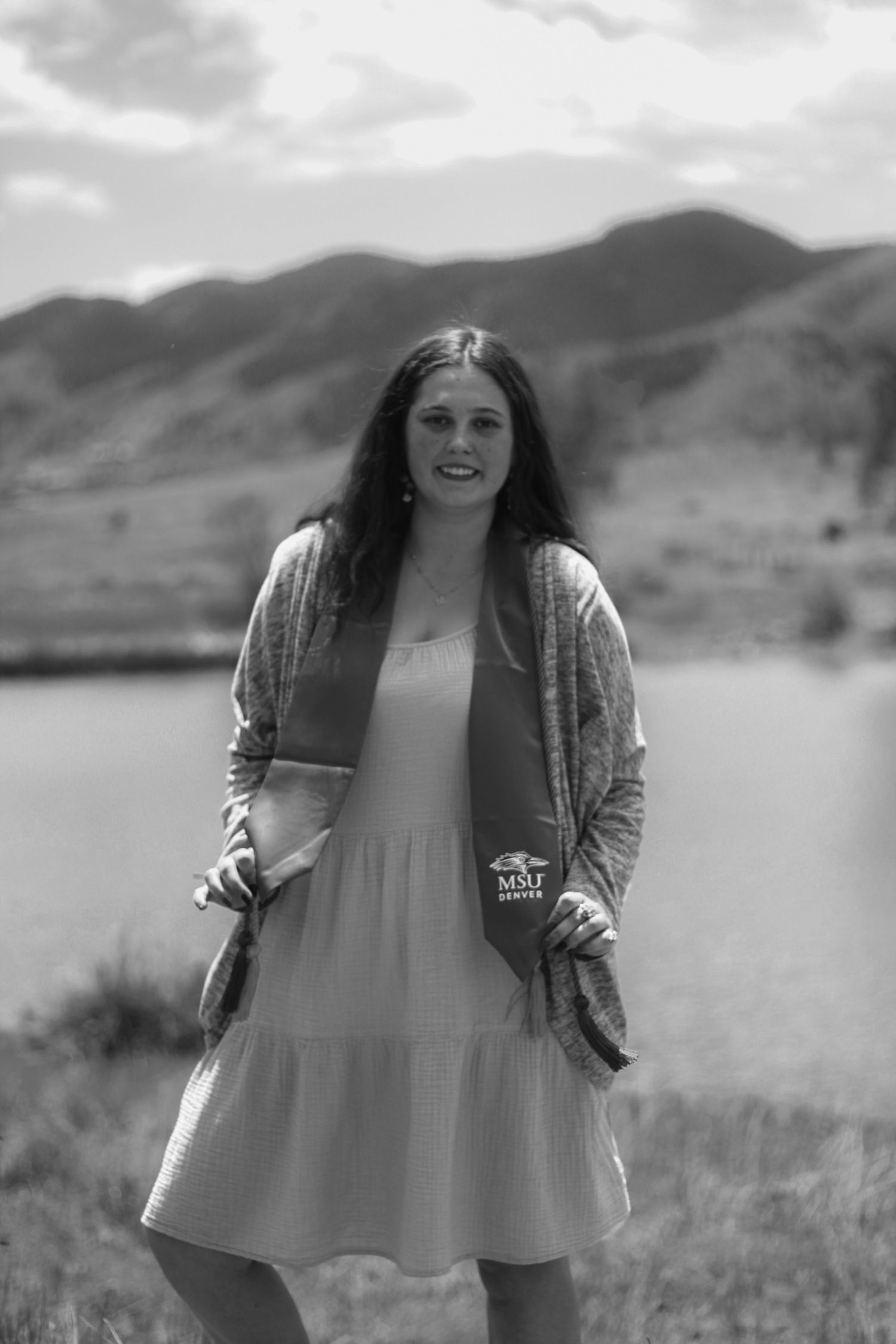 Black and white portrait of young woman in graduation stole standing by mountain lake.