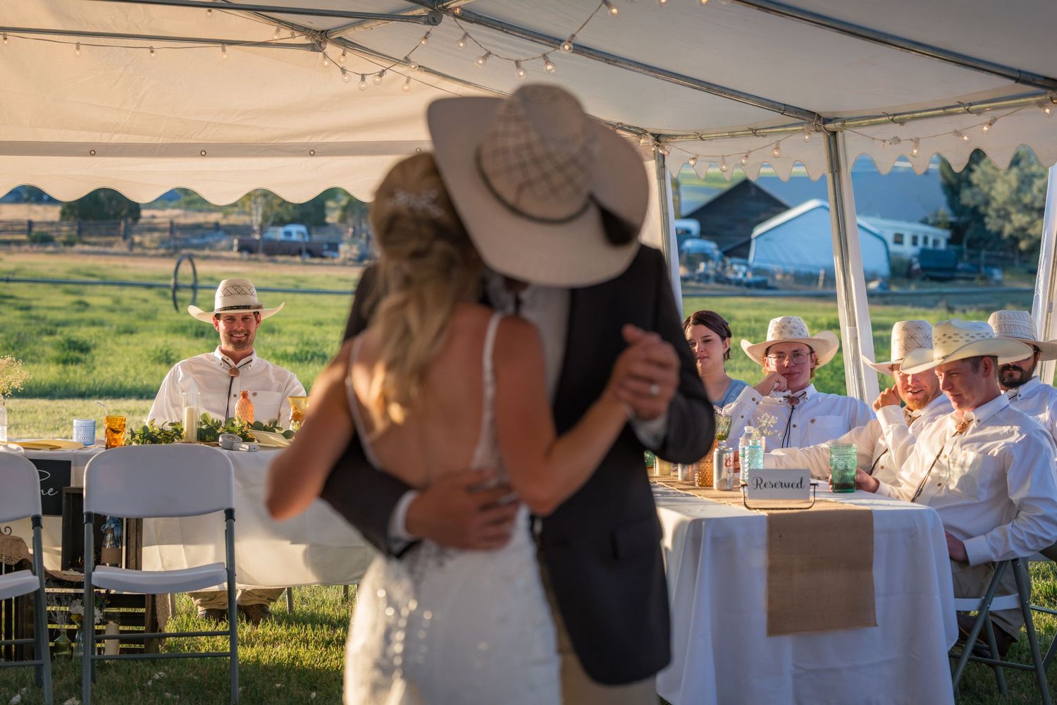 Couple sharing first dance at outdoor country wedding reception under tent with guests in cowboy hats seated at tables.
