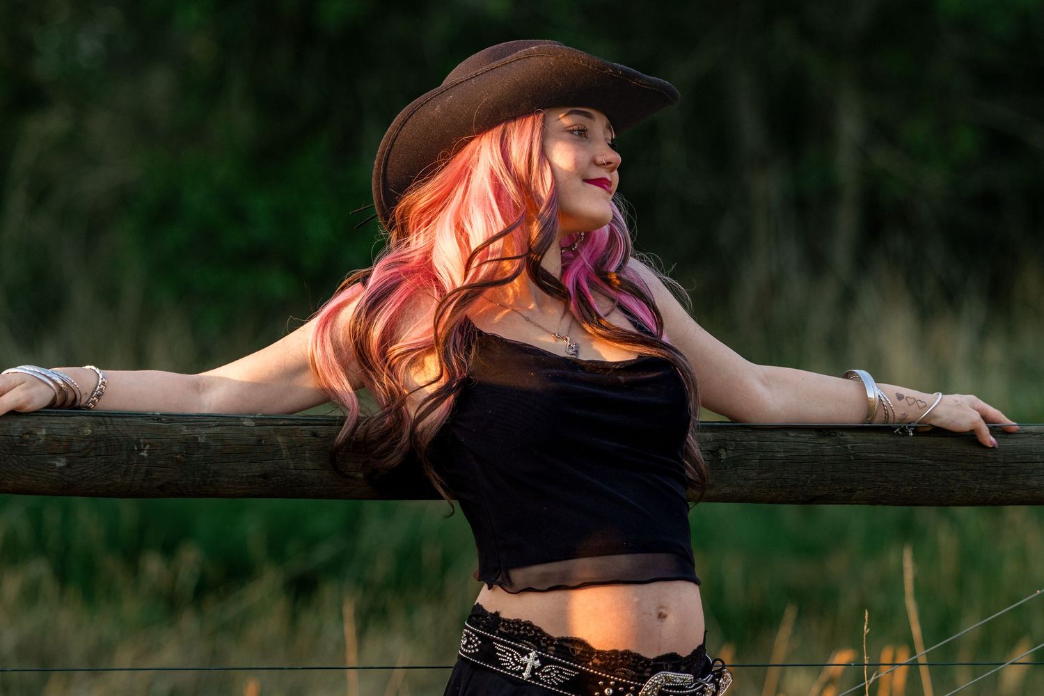 Young woman with pink hair wearing a cowboy hat leans against a wooden fence in a field.