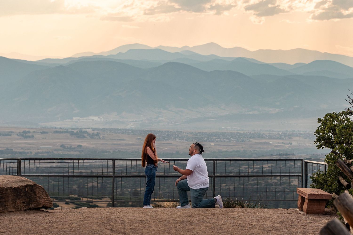 Man proposes to woman on scenic overlook with mountain sunset backdrop.