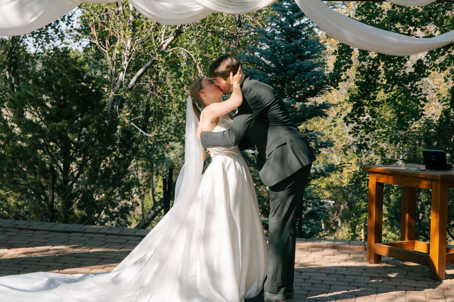Bride and groom share their first kiss at an outdoor wedding ceremony surrounded by lush green trees.