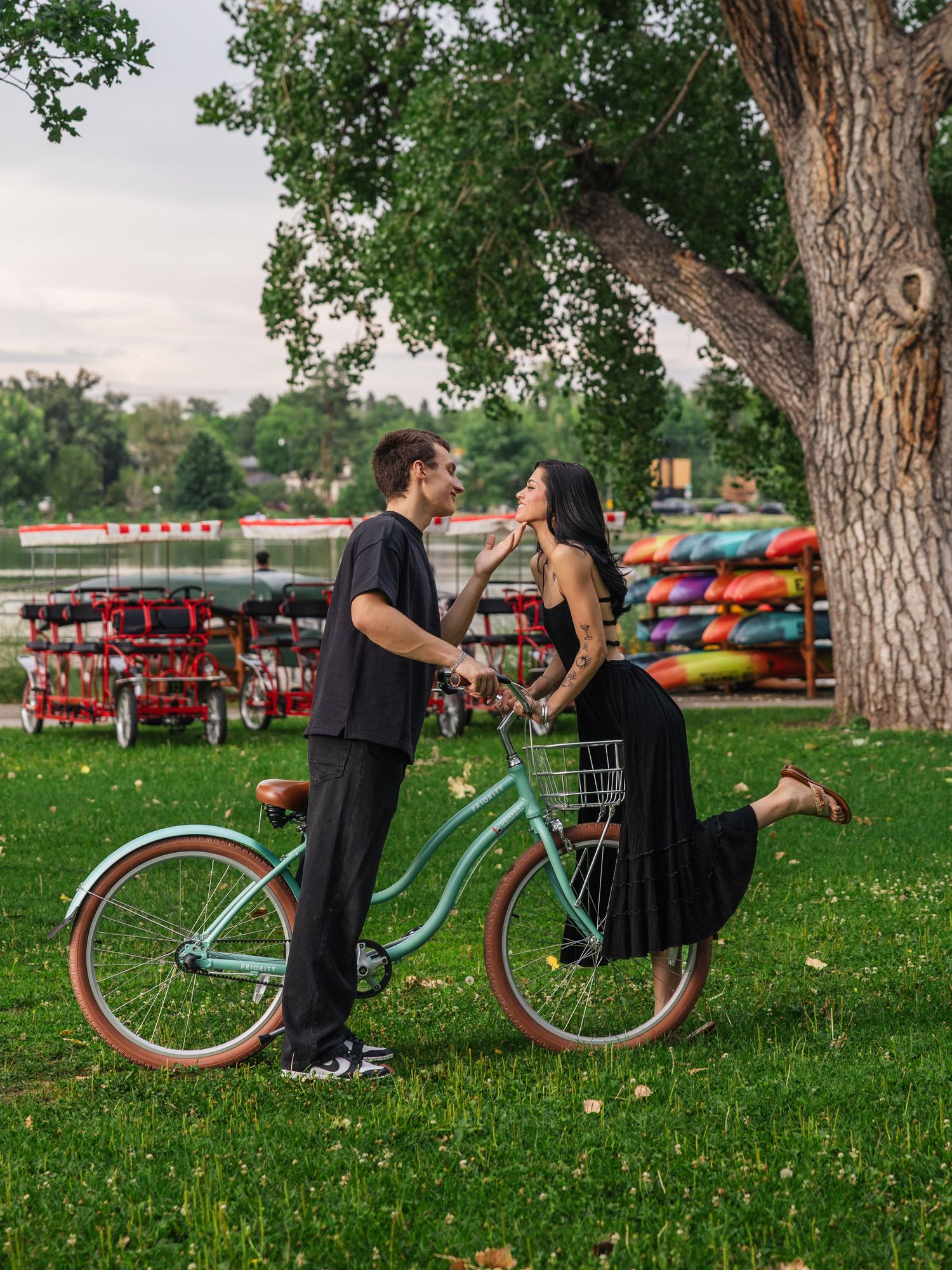 Couple sharing a romantic moment beside a teal vintage bicycle in a lush green park near a lake.