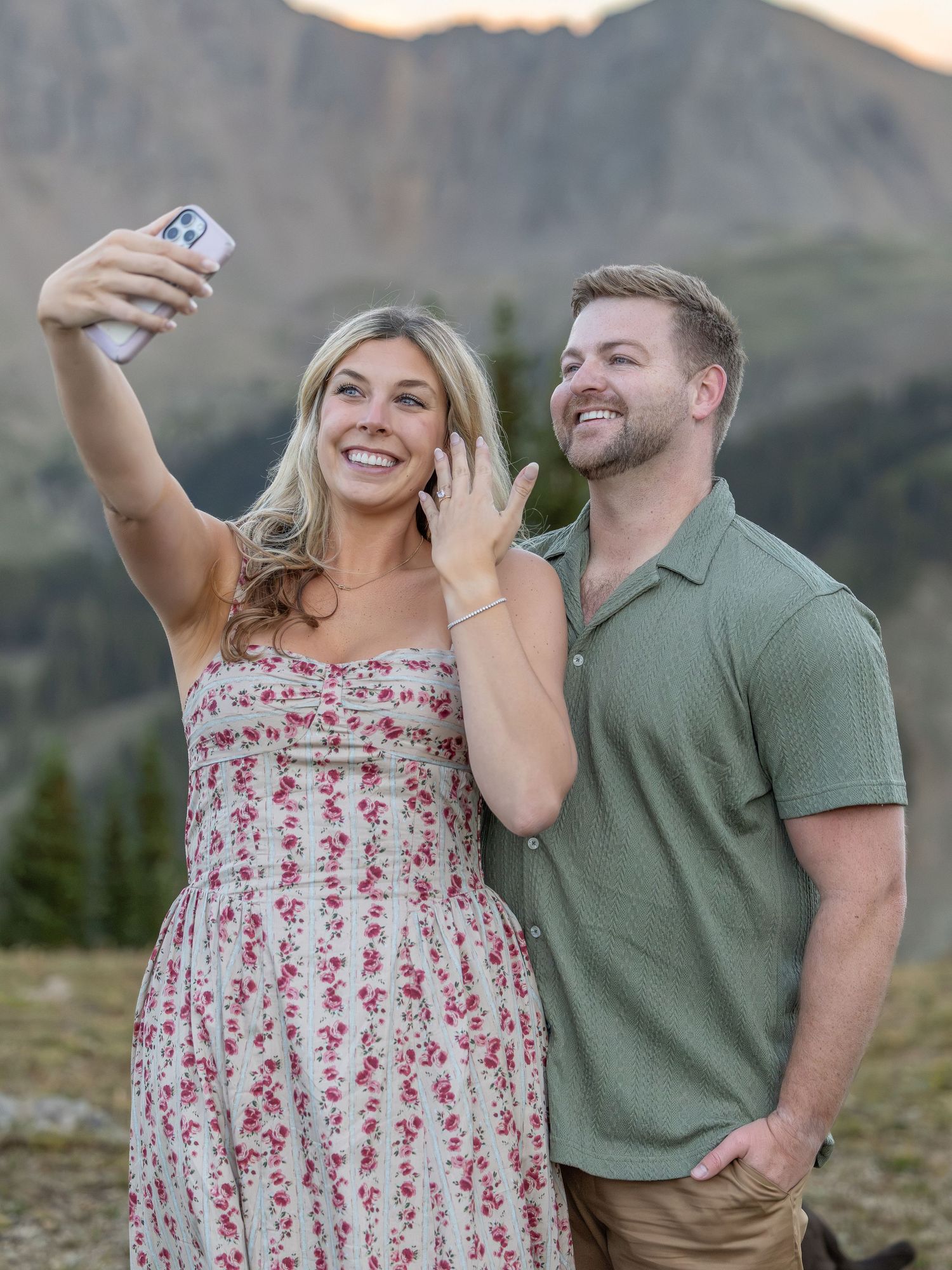A couple takes a selfie outdoors with mountain scenery, woman in floral dress showing engagement ring, man in green polo shirt.