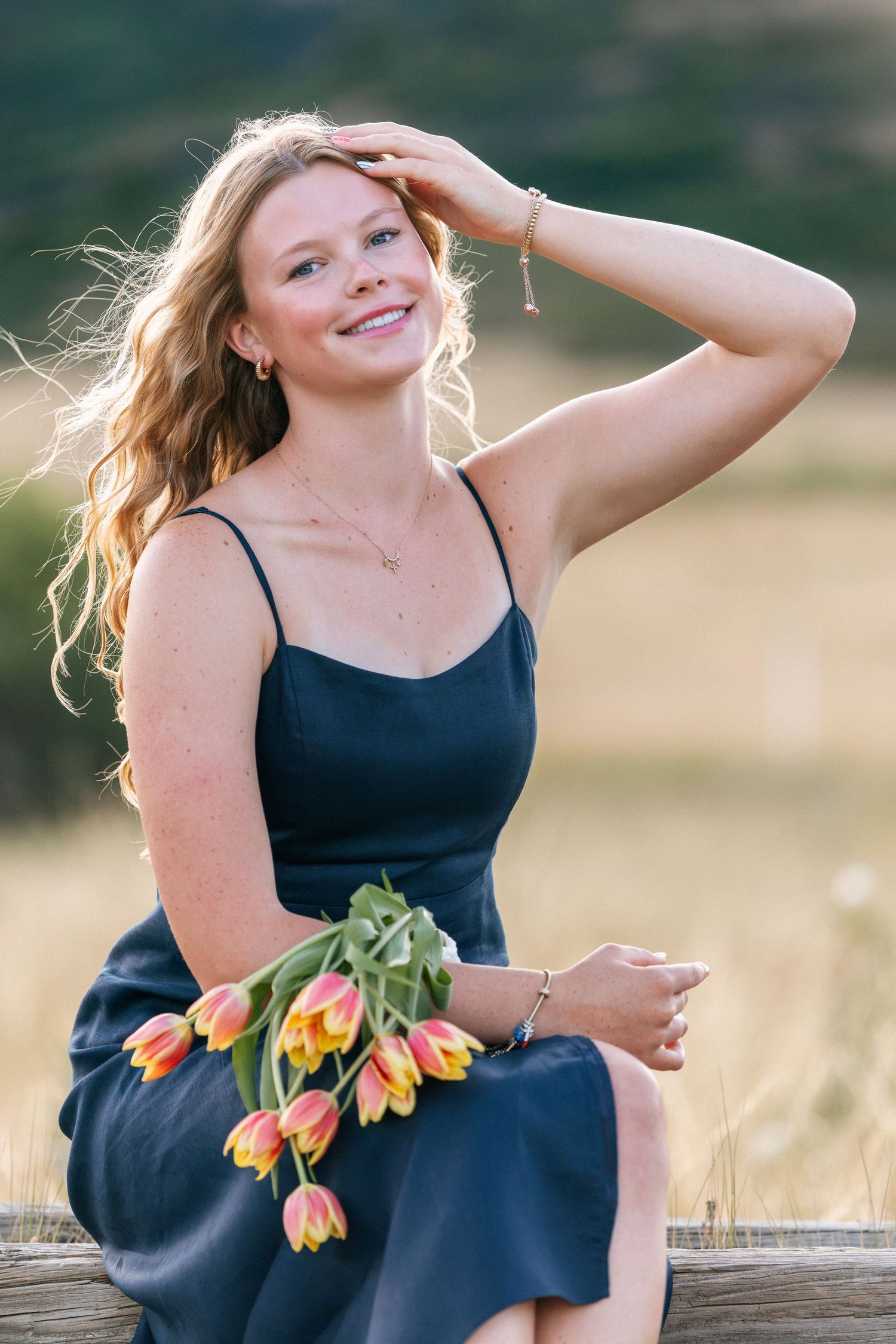 Young woman in navy dress holding tulips sits on fence, smiling in golden sunlight with hills in background.