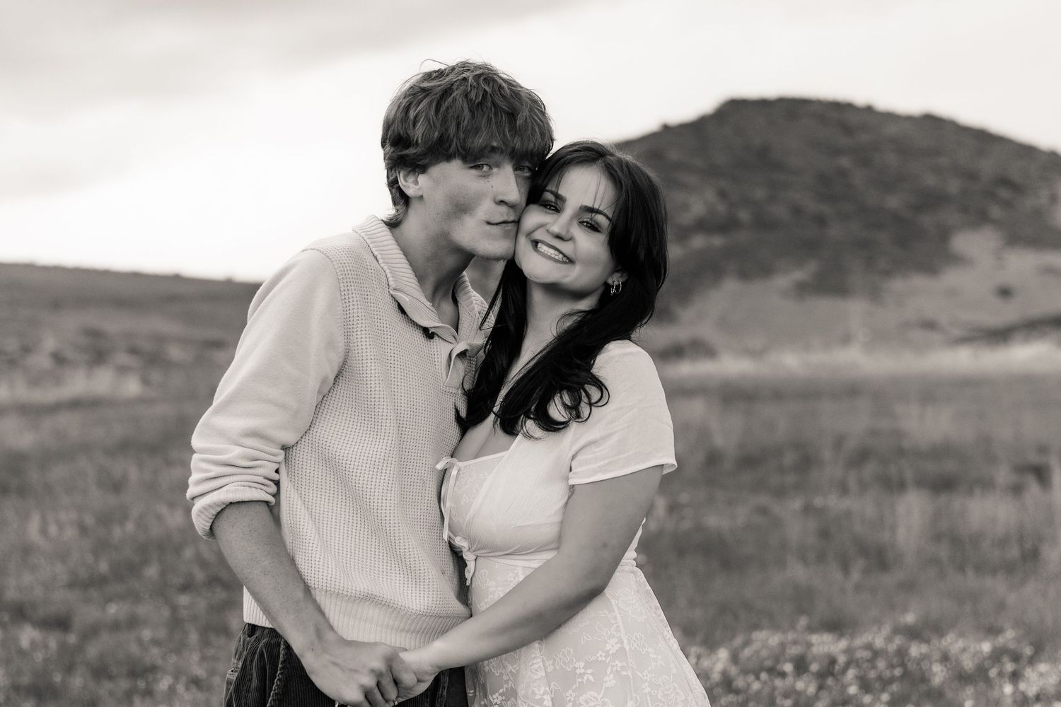 Black and white photo of a couple embracing and smiling in an open field with hills behind them in Littleton, Colorado.