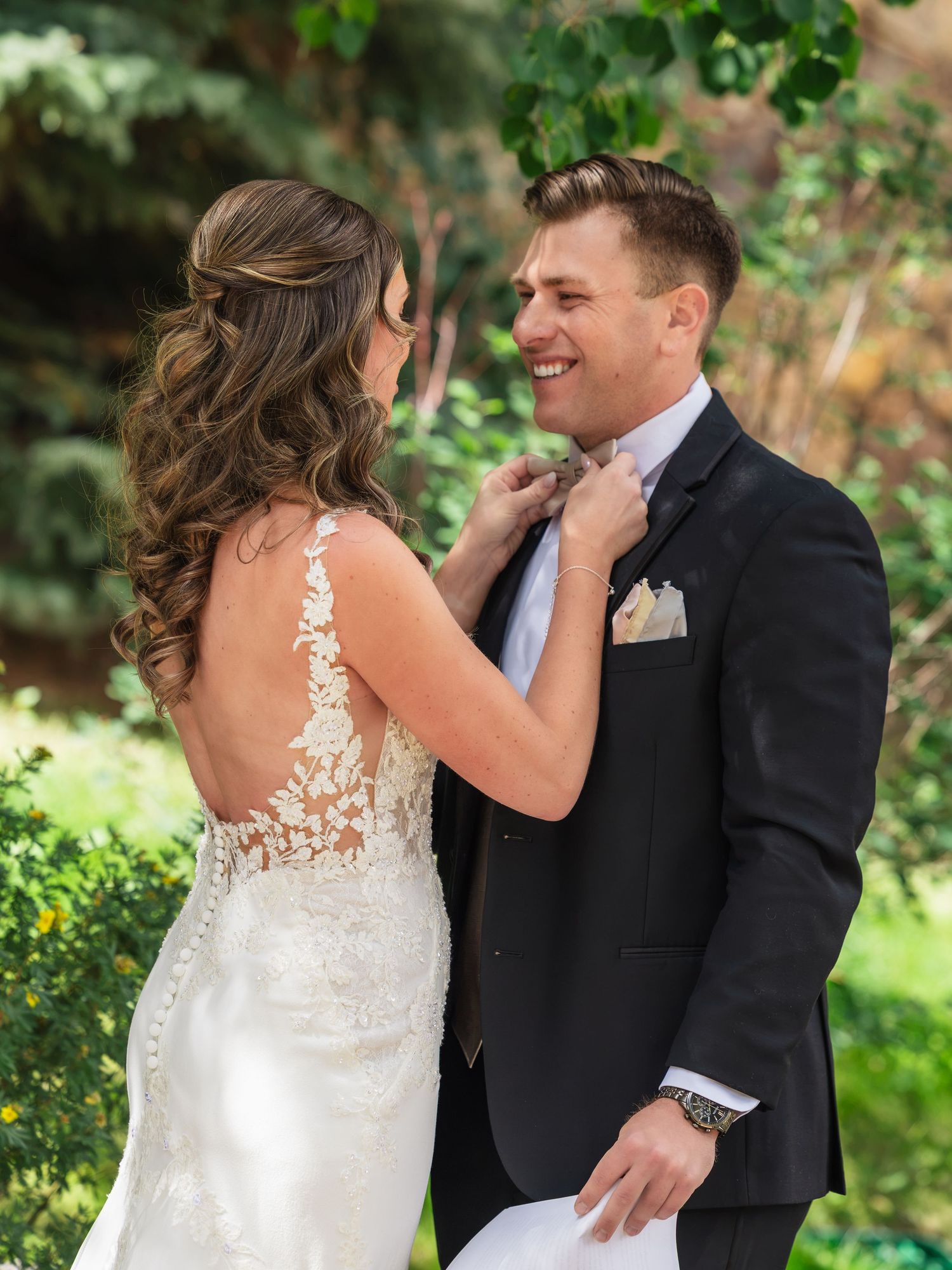 Bride in lace backless gown adjusts grooms black suit tie outdoors at wedding ceremony.