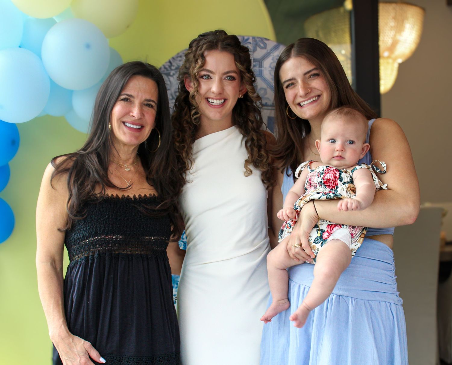Three women smiling together, one holding a baby girl, at a festive balloon-decorated celebration.
