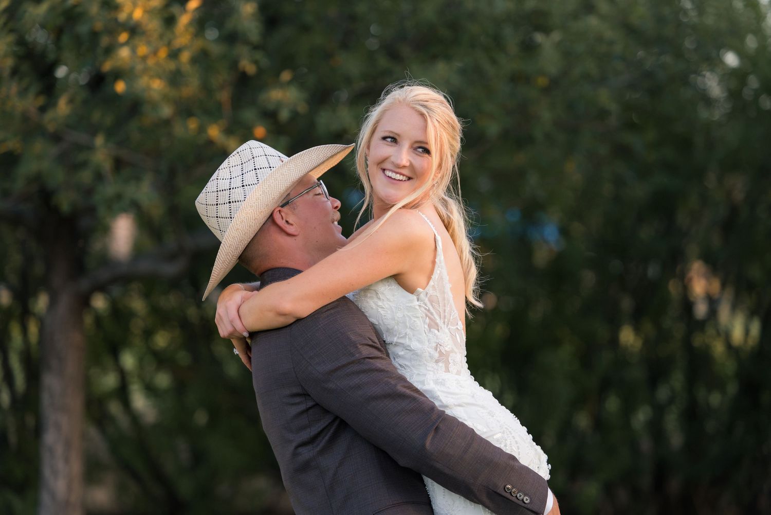 Groom in cowboy hat lifting smiling blonde bride in white lace dress outdoors at golden hour wedding.