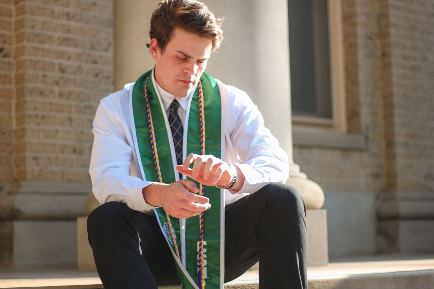 Young man in white shirt and green graduation stole sits on steps looking at his hands.