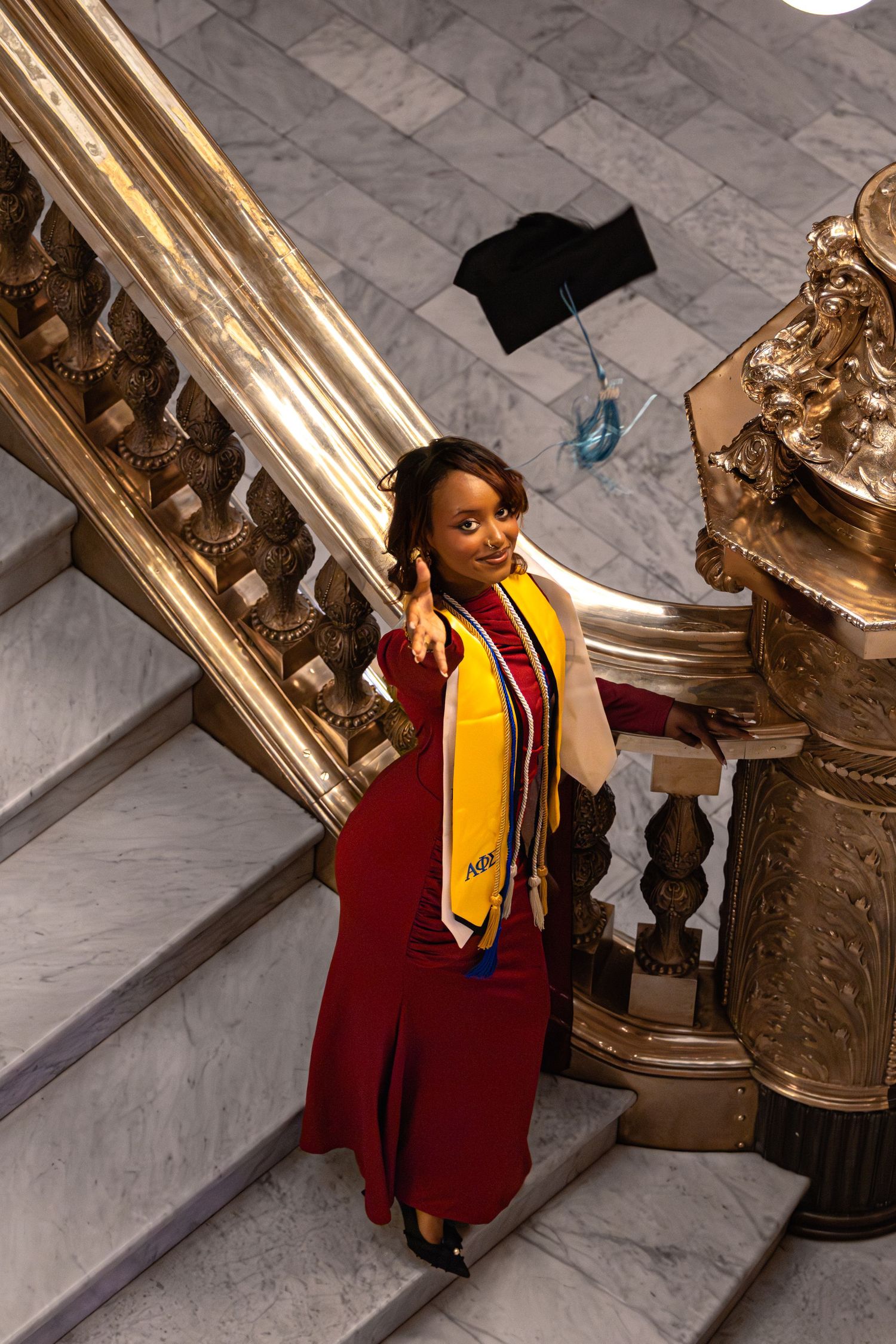 A smiling graduate in red regalia and gold stole poses on ornate marble stairs, with a mortarboard tossed in the air.