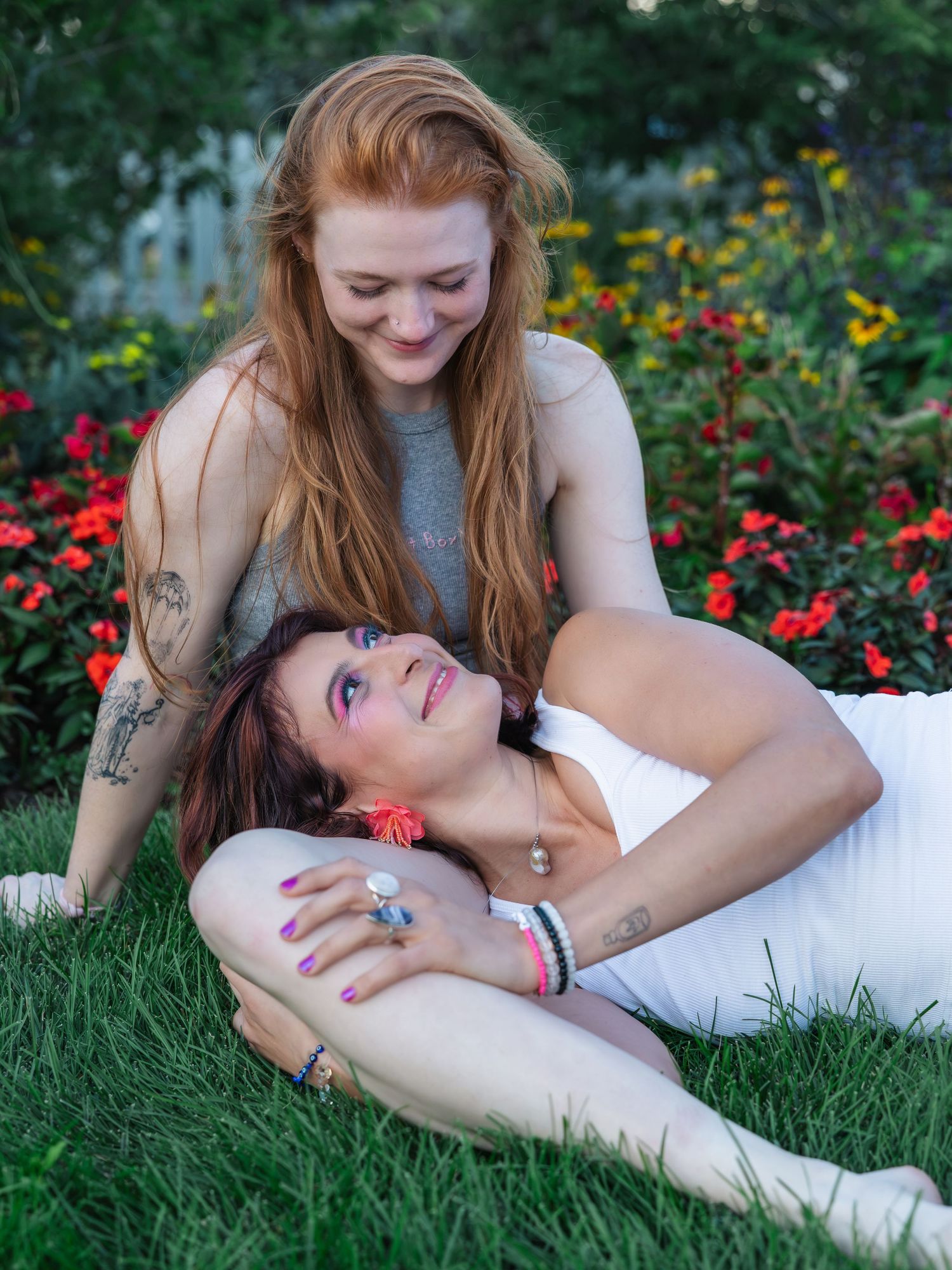 Two women relaxing together on green grass surrounded by colorful flowers in a garden setting.