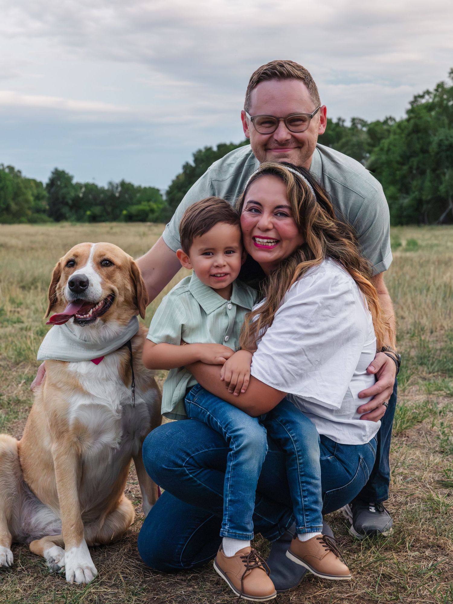 Family of three with a beagle dog posing outdoors in a field, smiling together during a portrait session.