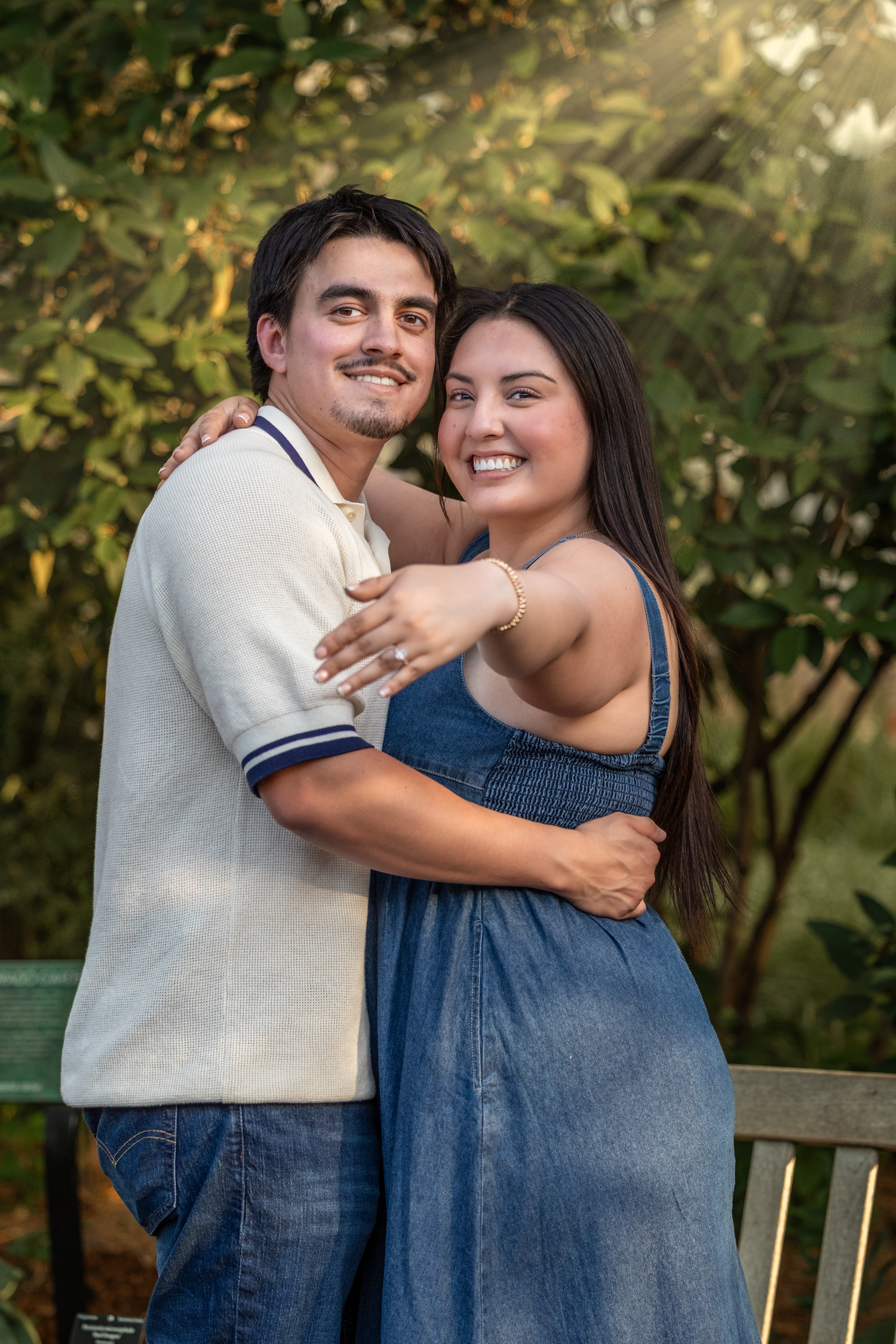 A smiling couple embraces outdoors, woman in denim dress and man in white polo, with lush green trees in background.