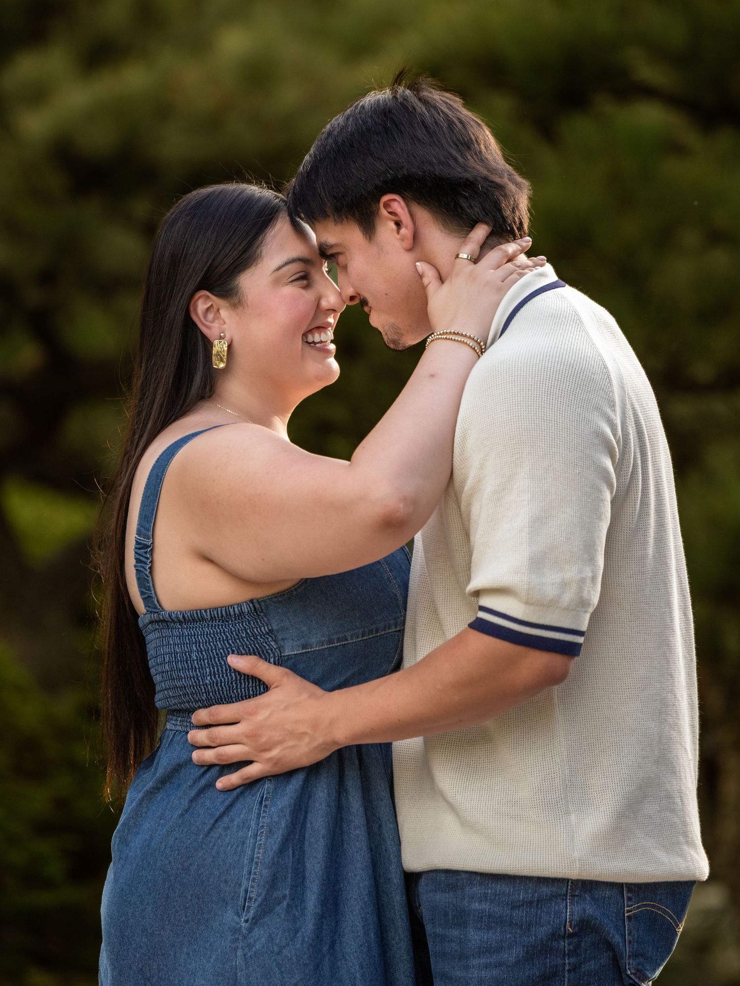 A happy couple shares a romantic moment outdoors, smiling and embracing near lush green trees.