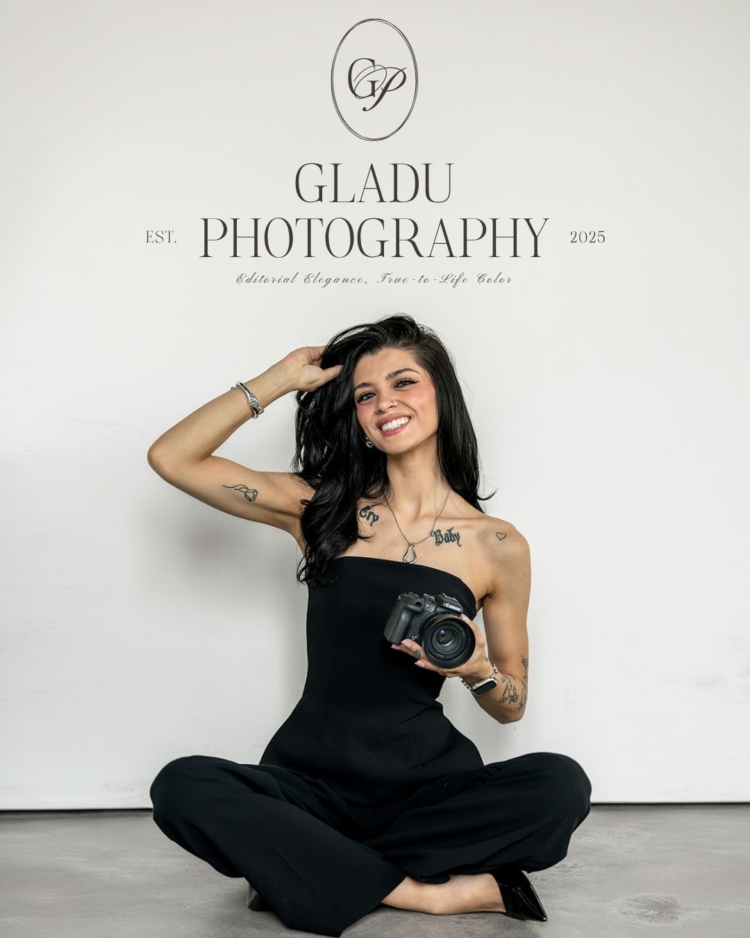 Woman photographer sitting cross-legged holding camera, smiling in front of Gladu Photography studio backdrop.