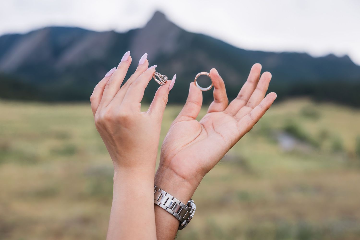 Two hands holding wedding rings outdoors with mountains and green fields in the background.