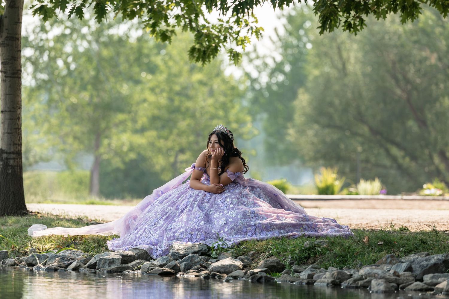 Young woman in lavish lilac ball gown posing by a serene pond in a lush green park setting.