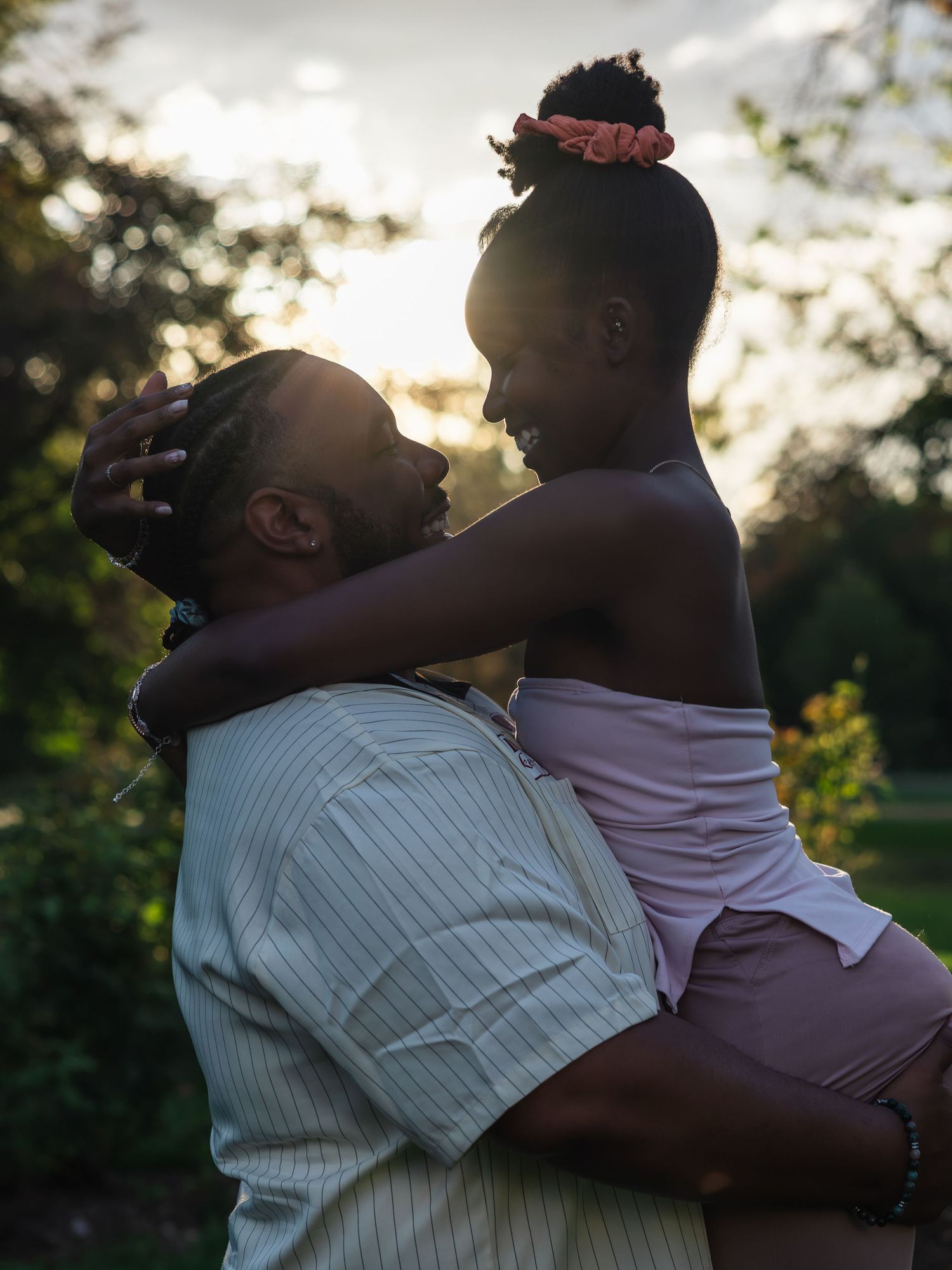 A romantic couple embraces at golden hour, the man lifting a woman in pink as they gaze at each other outdoors.