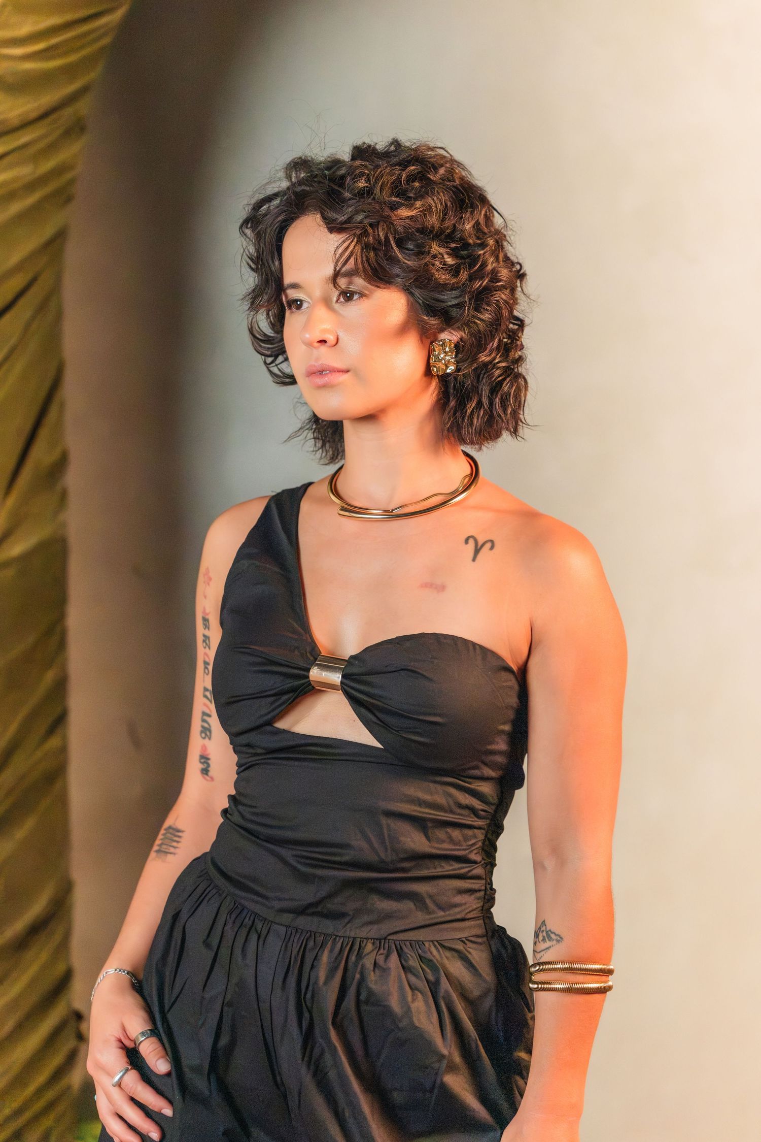 Woman in black one-shoulder dress with gold jewelry and tattoos, posing confidently indoors.