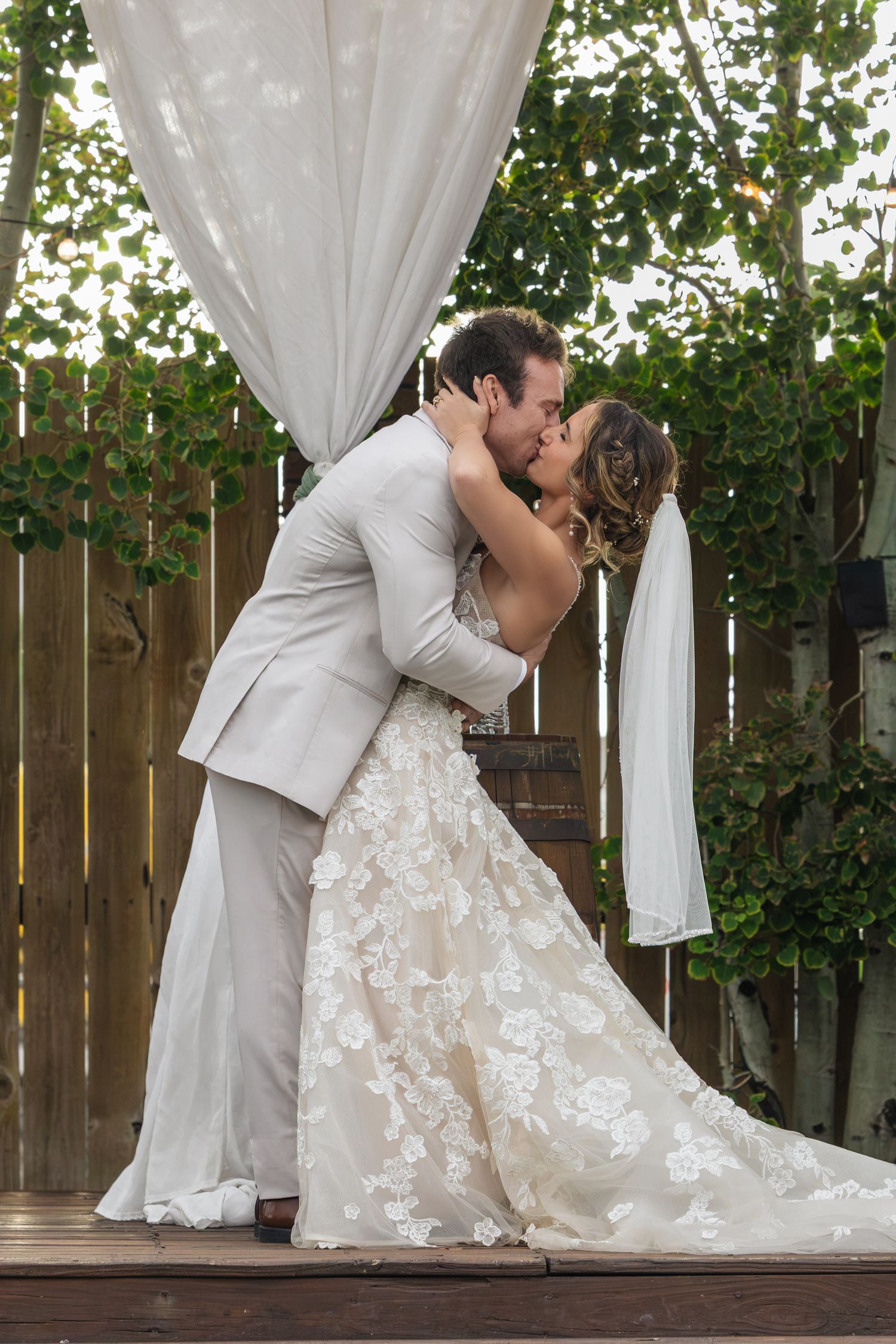 Bride and groom share first kiss at outdoor wedding ceremony beneath white draped arch surrounded by greenery.
