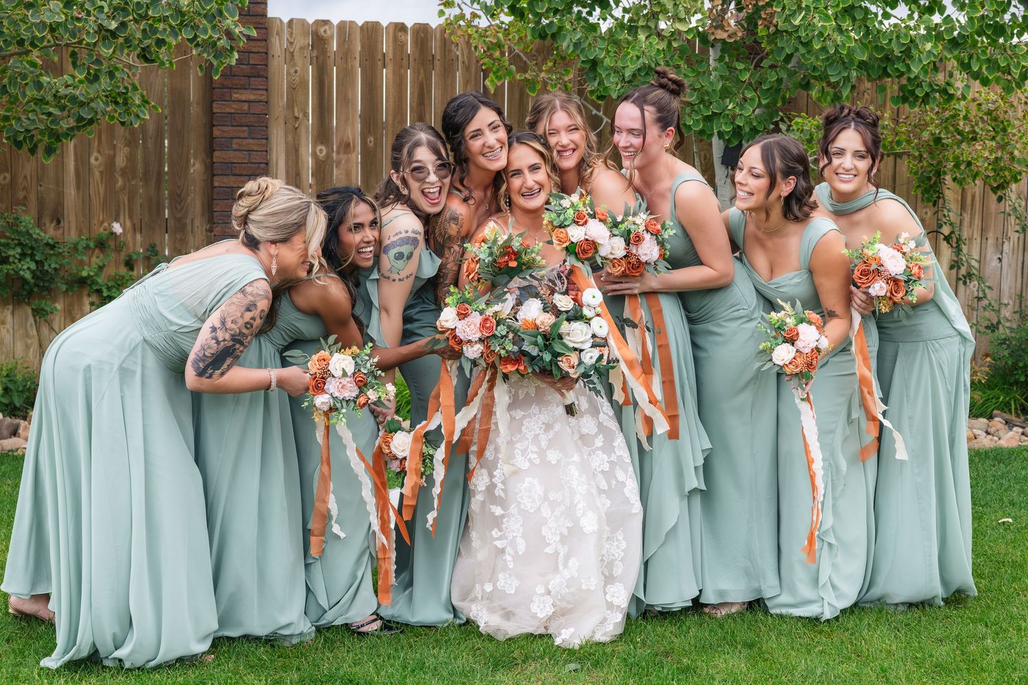 Bride in floral gown poses with bridesmaids in sage green dresses holding orange and white bouquets with ribbon streamers.