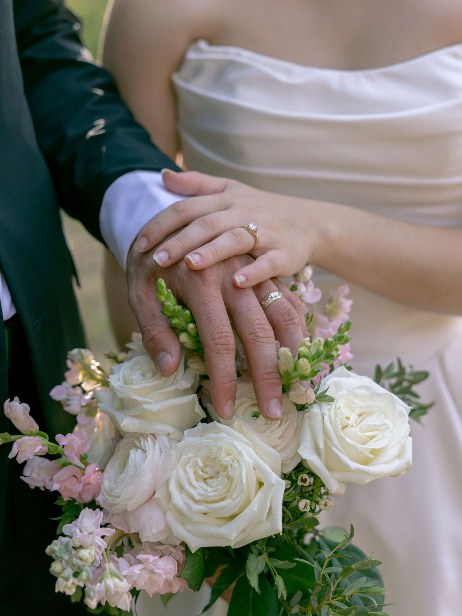 Newlyweds display wedding rings on clasped hands resting over a bridal bouquet of white roses and pink flowers.