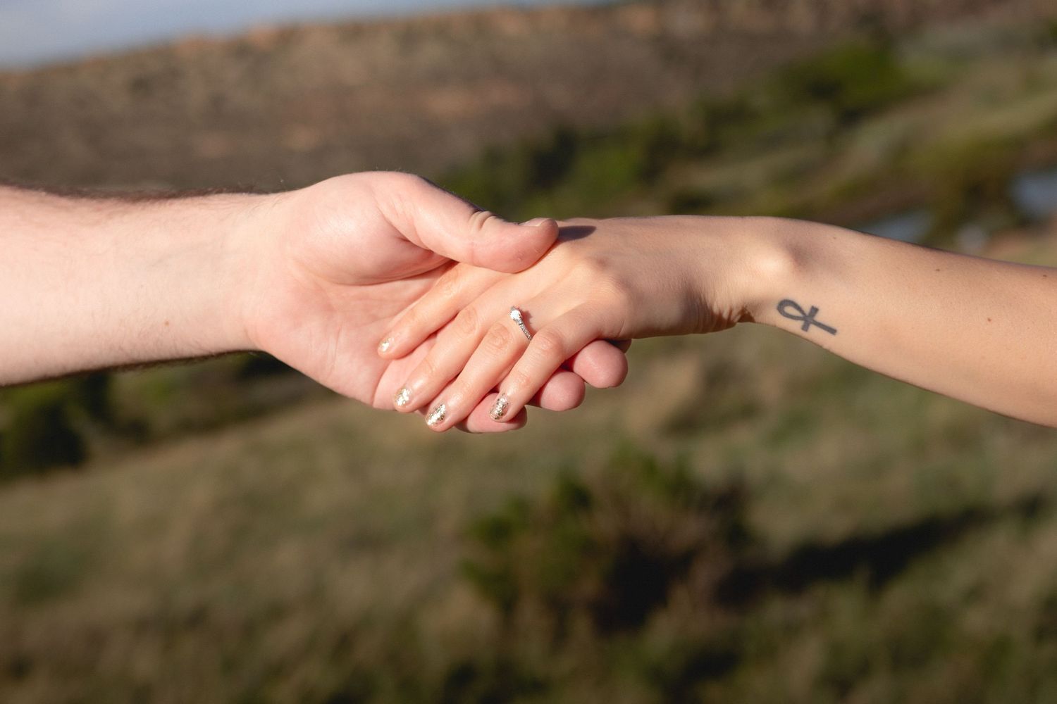 Two people holding hands outdoors, one with an ankh tattoo on their wrist, with greenery in the background.