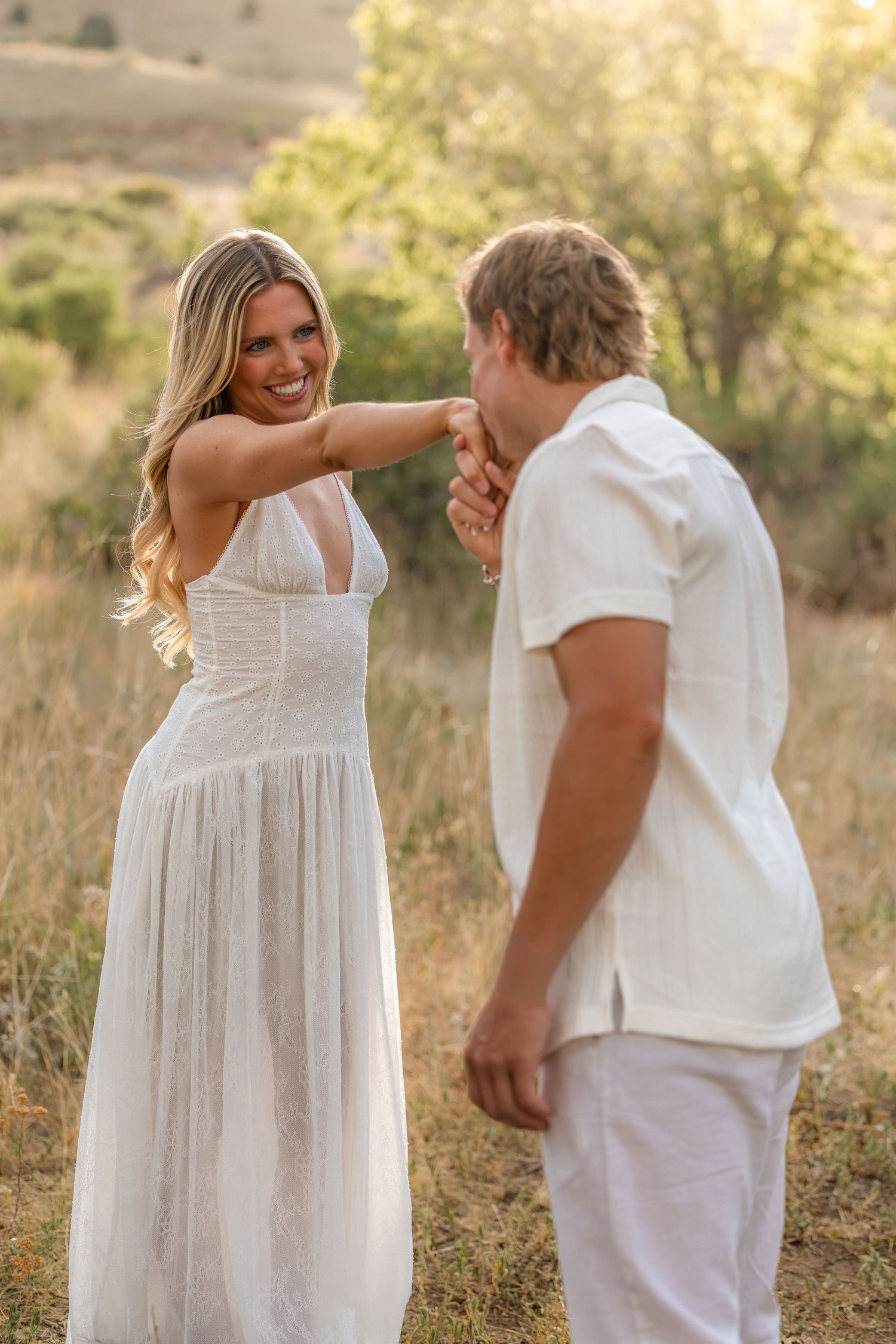 Smiling blonde woman in white dress having hand kissed by man in white outfit in golden sunlit field.
