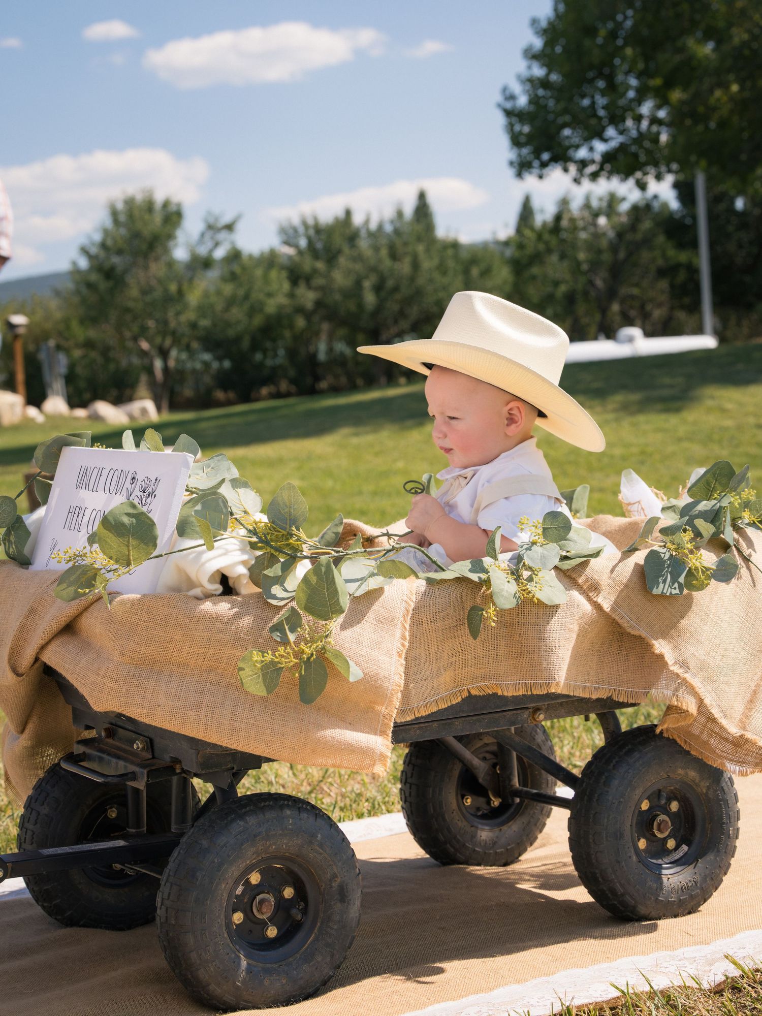 A cute baby in a cowboy hat rides in a burlap-decorated wagon adorned with greenery at an outdoor wedding.