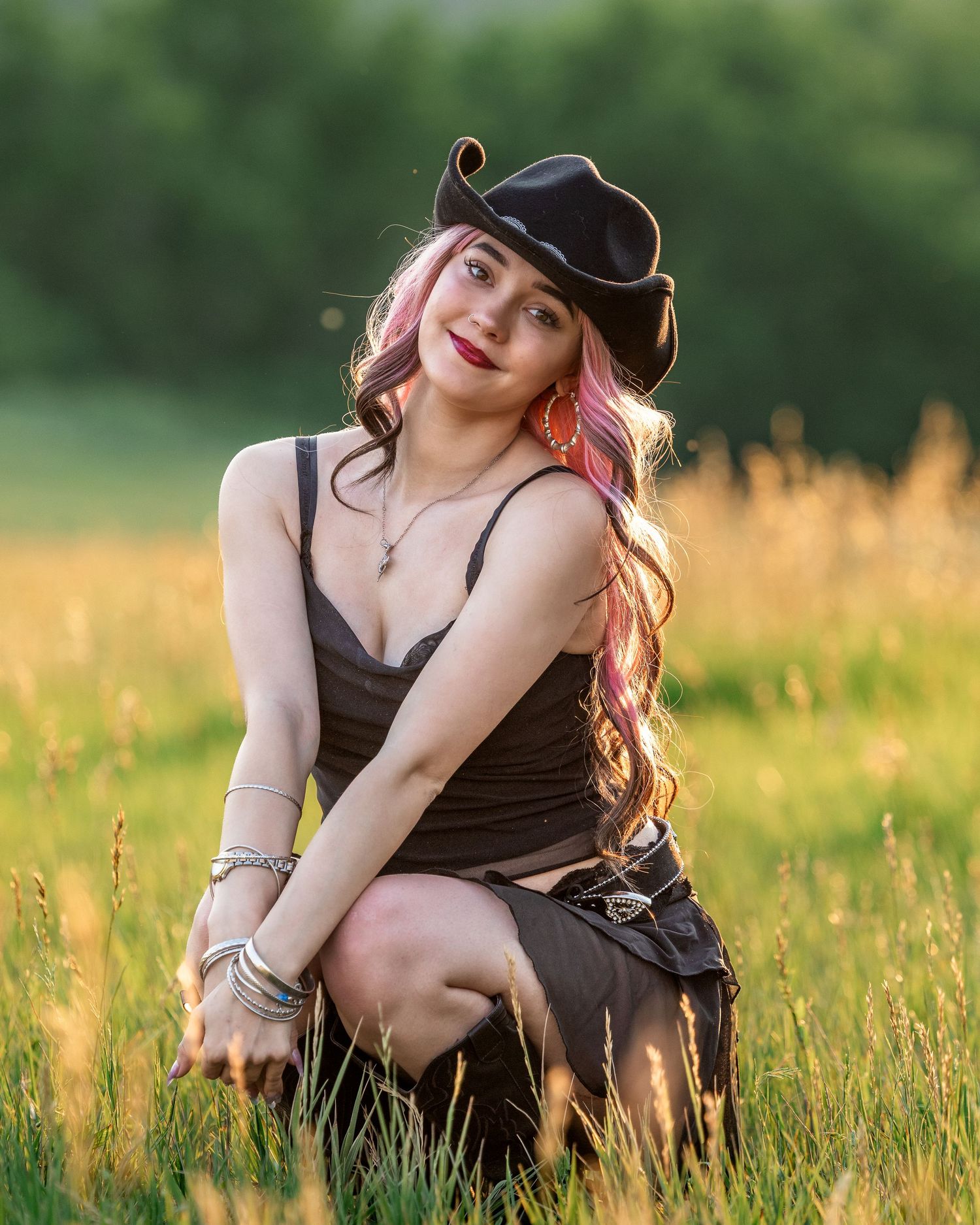 Young woman with pink hair wearing a black cowboy hat and dark dress poses in a sunlit grassy field.