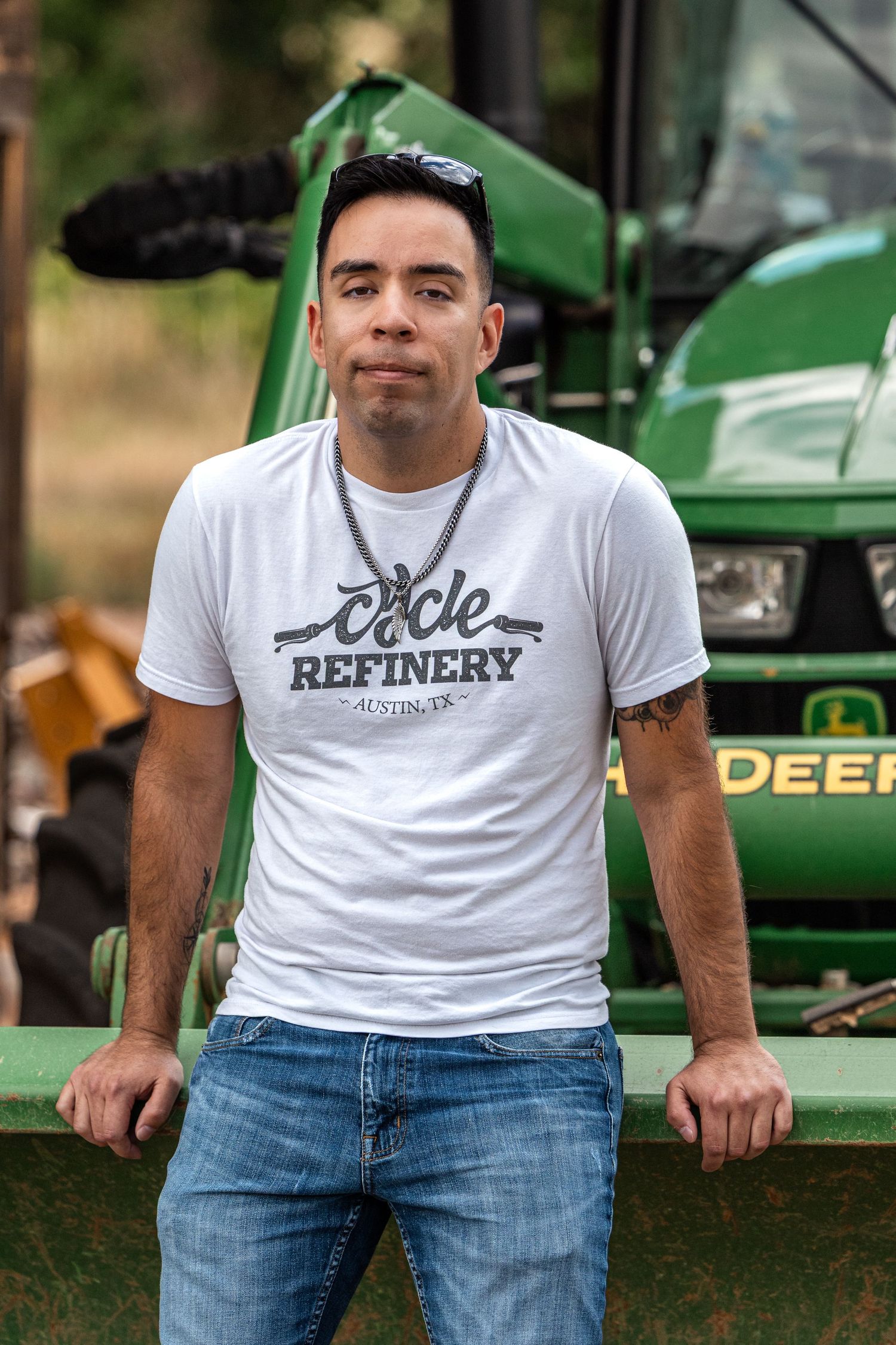 Man in white Circle Refinery Austin TX t-shirt leaning against green John Deere tractor outdoors.