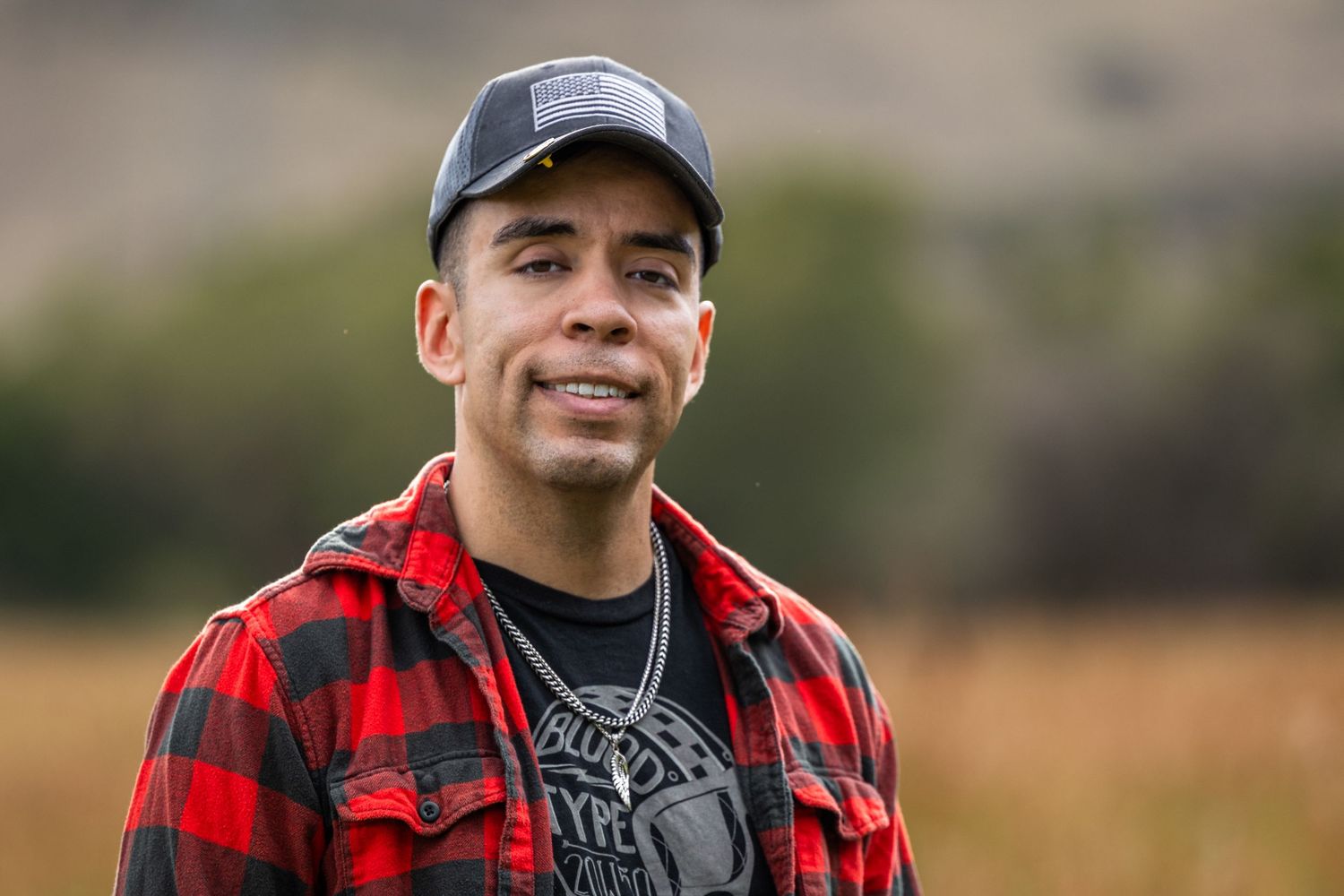 Man in red flannel shirt and gray cap smiling outdoors in a green field.