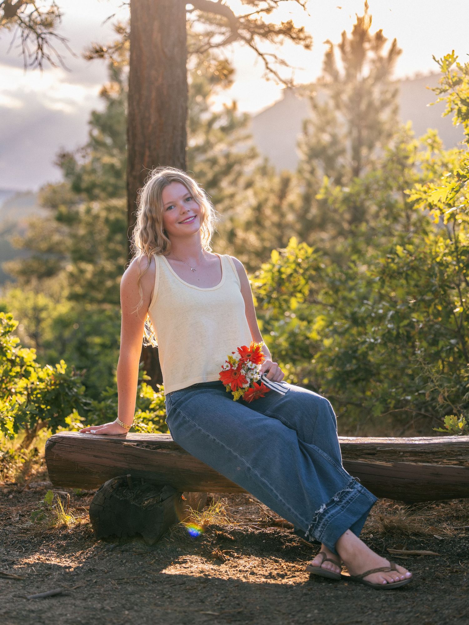 Young woman in jeans and cream top sits on log bench holding red flowers with mountain sunset backdrop.