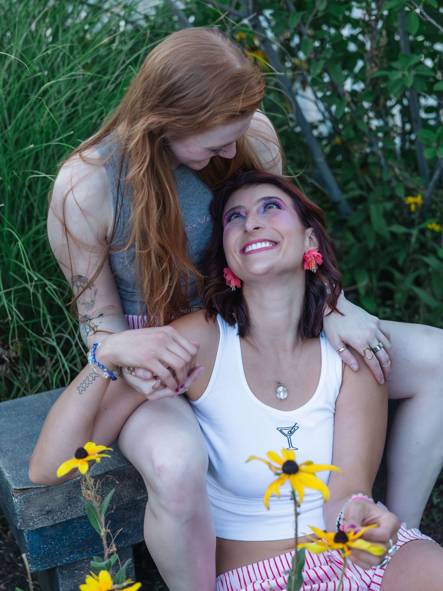 Two women smile and hold hands outdoors among yellow black-eyed Susan flowers, one with red hair and tattoos, one in a white top.