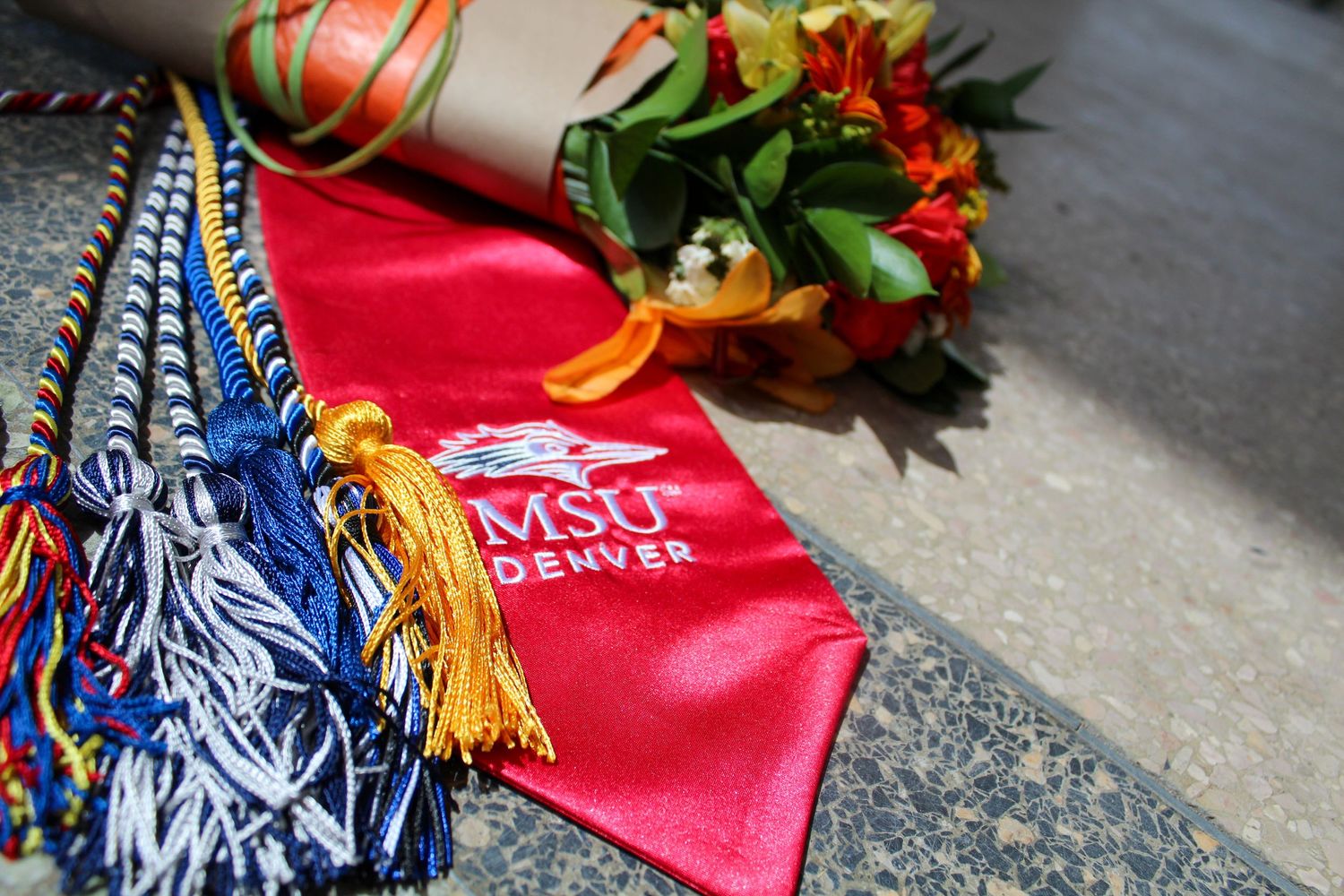 MSU Denver graduation regalia with red sash, colorful honor cords, and a bouquet of flowers on a stone surface.