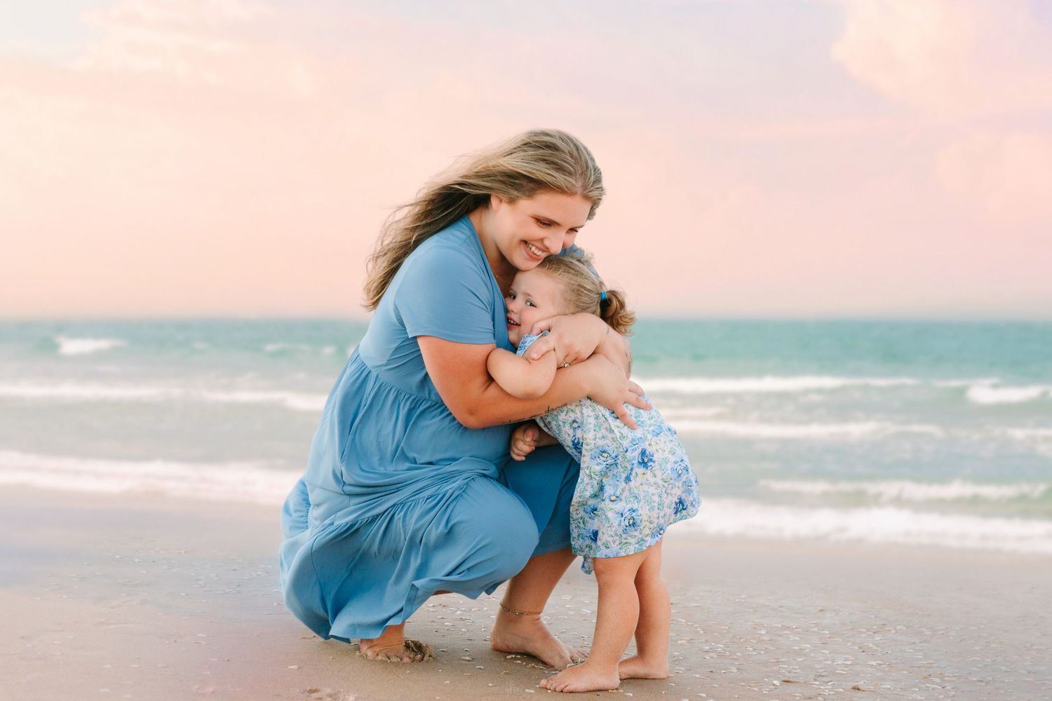 A mother in a blue dress hugs her toddler daughter on a sandy beach at sunset.