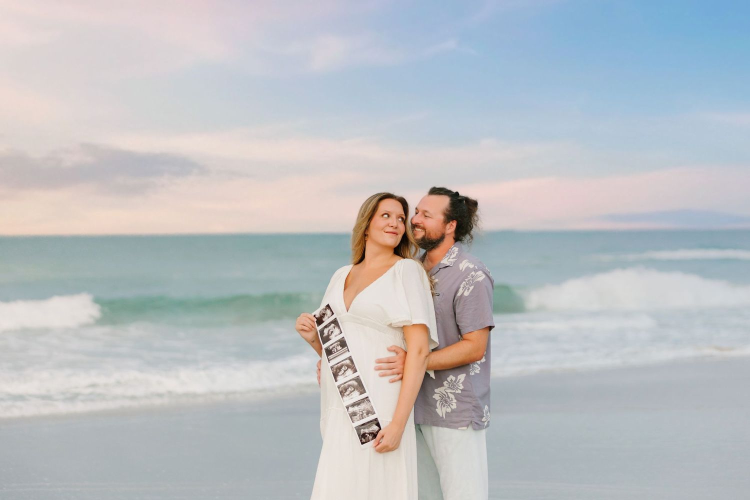 Pregnant couple embracing on a beach at sunset, woman in white dress holding ultrasound photos with a mom-to-be sash.