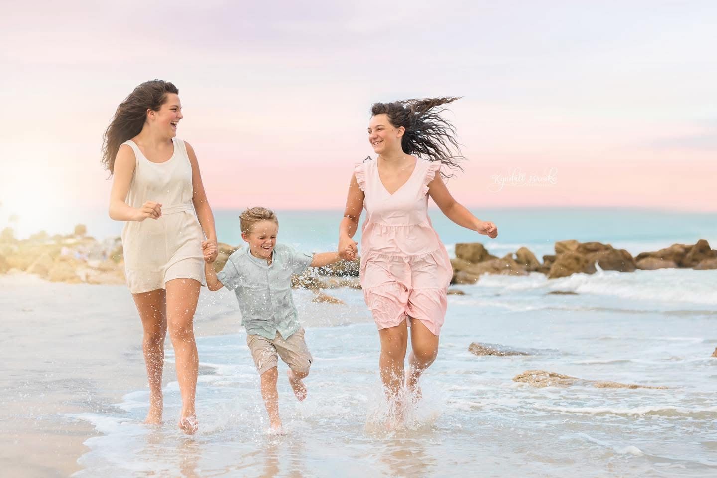 Two women and a child joyfully run along a sunny beach shoreline, splashing in the waves together.