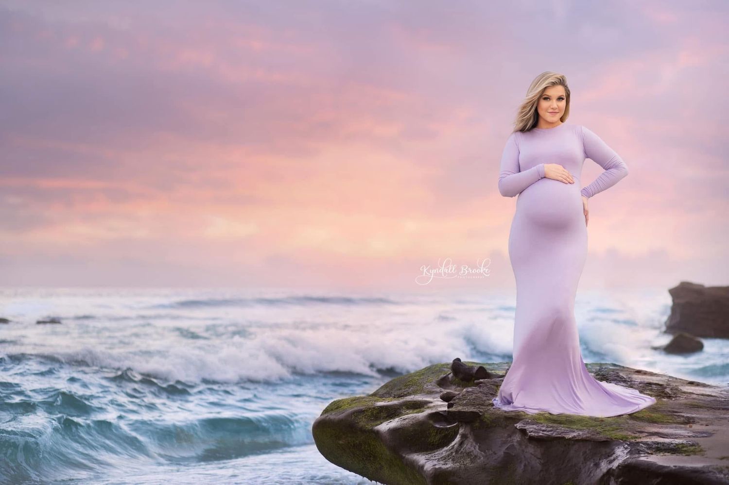 Pregnant woman in lavender gown standing on coastal rocks at sunset with dramatic ocean waves and pink sky.