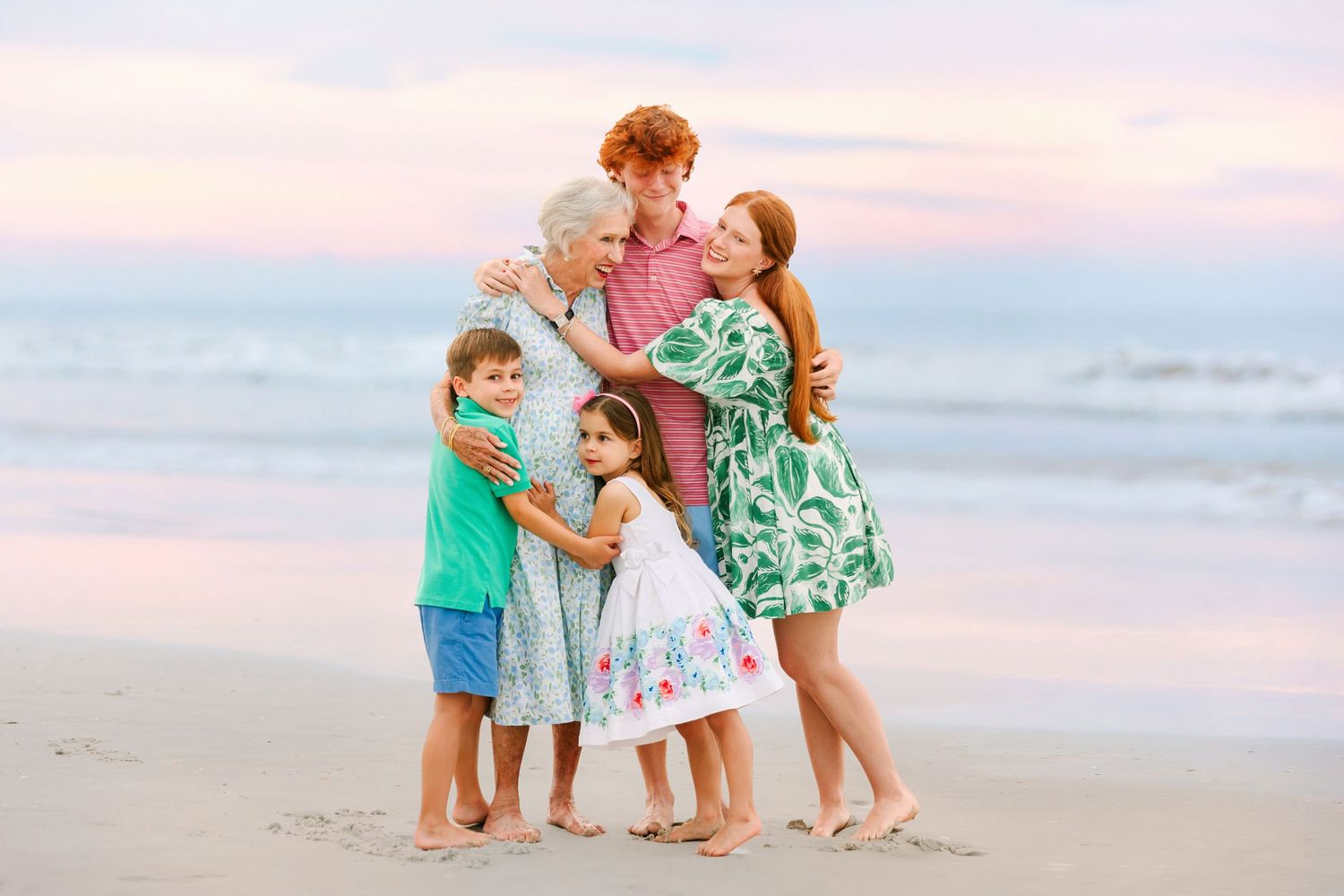 Family of five with redheads embracing on a beach at sunset, wearing pastel outfits.