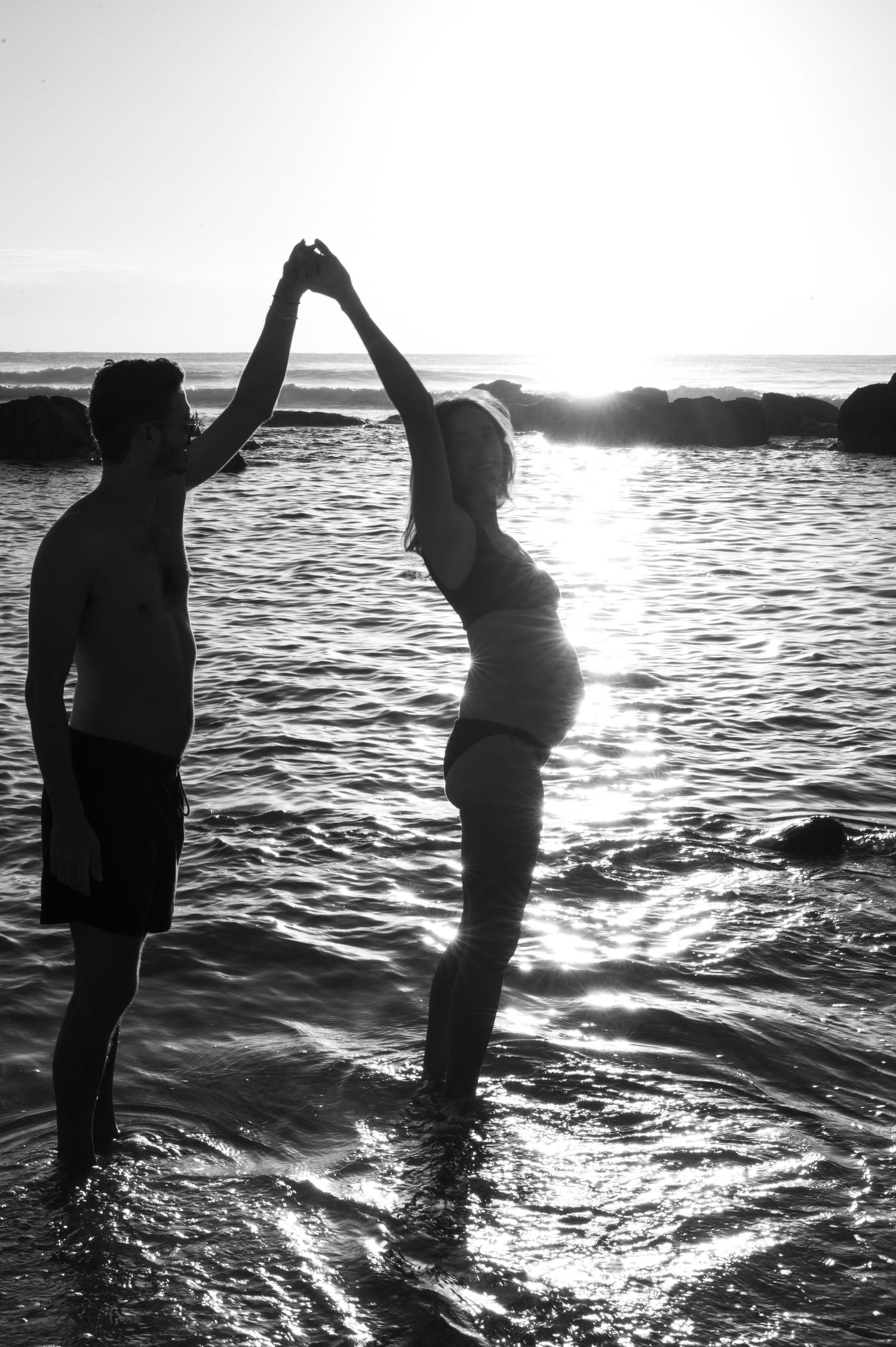 Couple holding hands dancing in ocean waves at sunset, black and white beach photography. Maternity Photography
