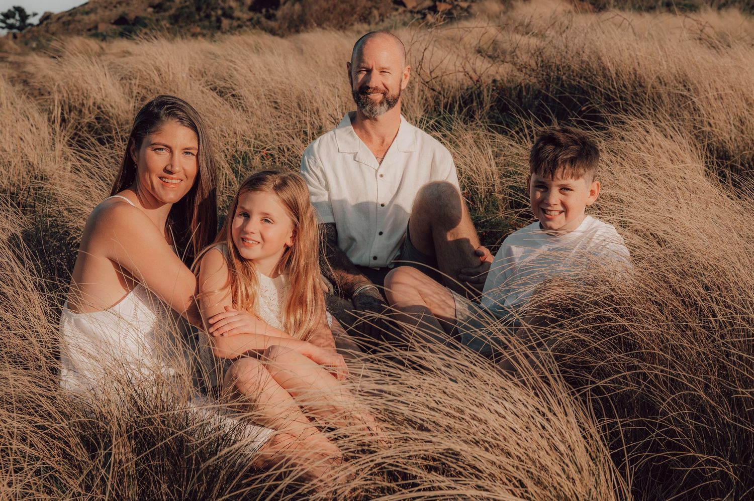 Family of four smiling together in golden grass field, wearing white outfits during a warm outdoor photo session. Family Photography