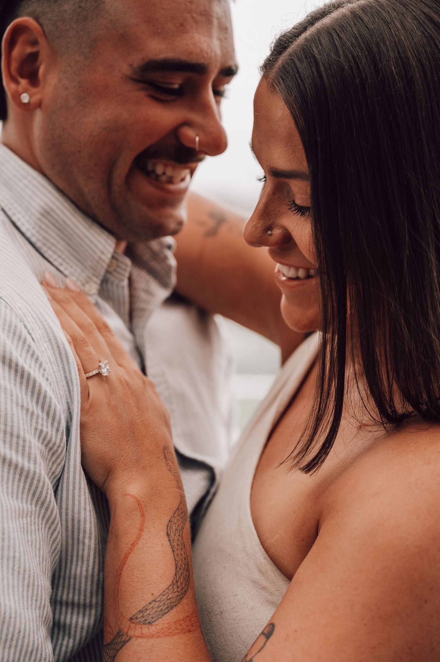 Smiling couple sharing a close romantic moment, woman displaying engagement ring and tattoos on her arm. Proposal. Proposal Photography