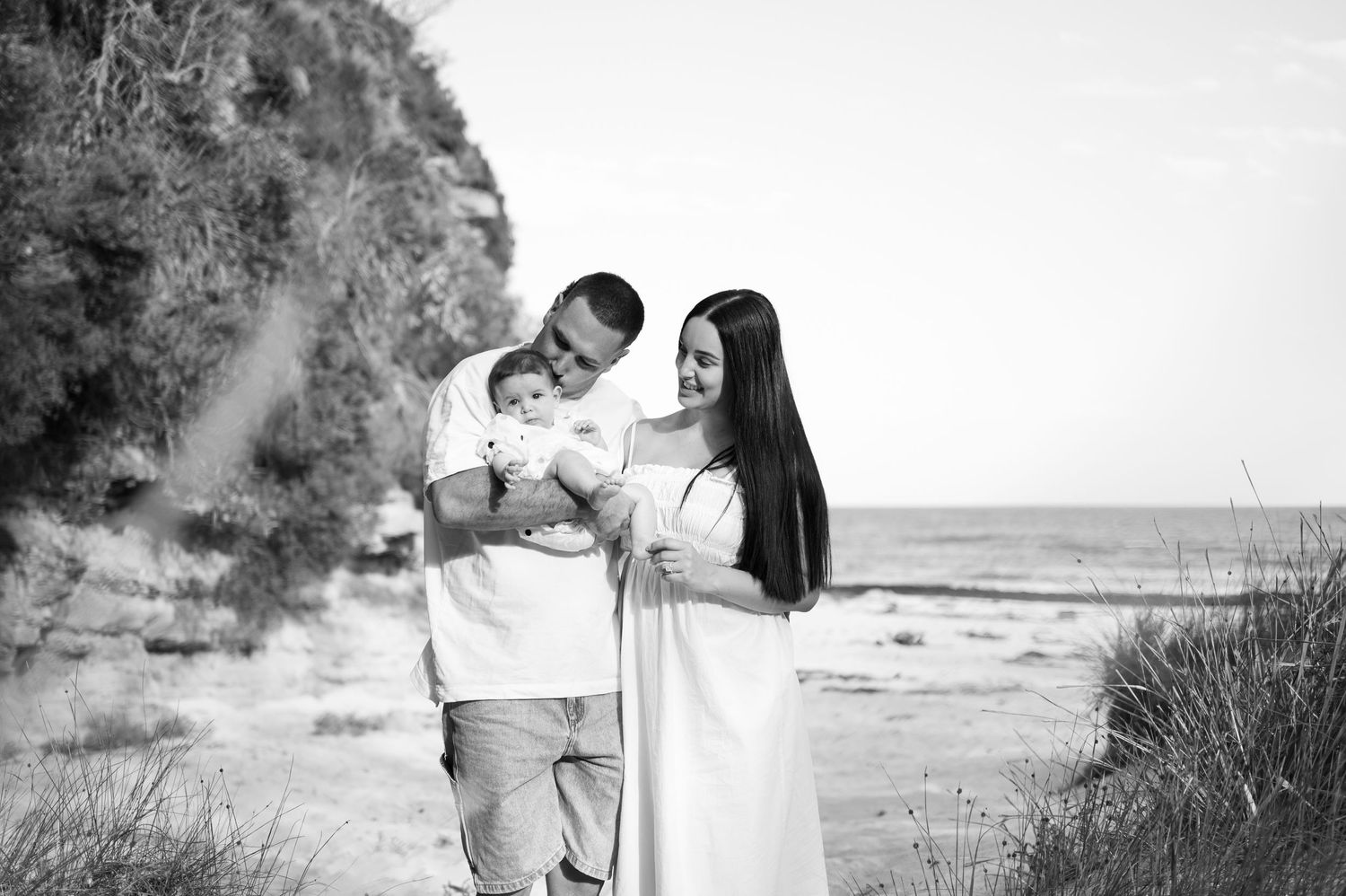 Black and white photo of a couple on a beach holding their newborn baby, with trees and ocean in the background.
Family Photography