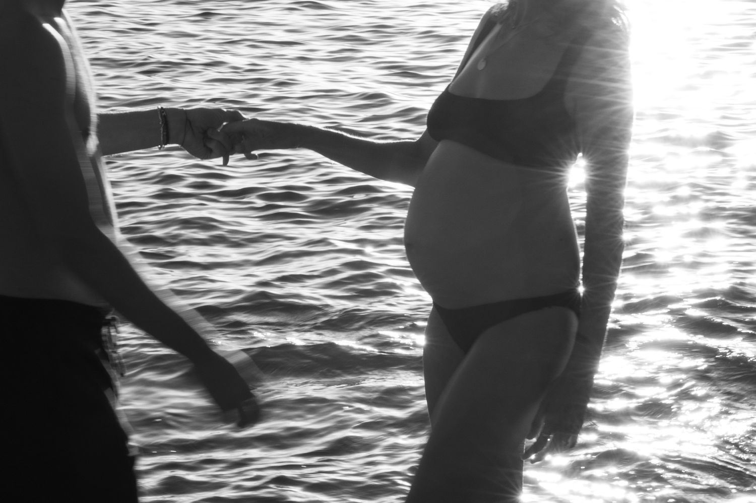 Black and white photo of a pregnant woman in a bikini holding hands with a partner near the ocean. Maternity Photography