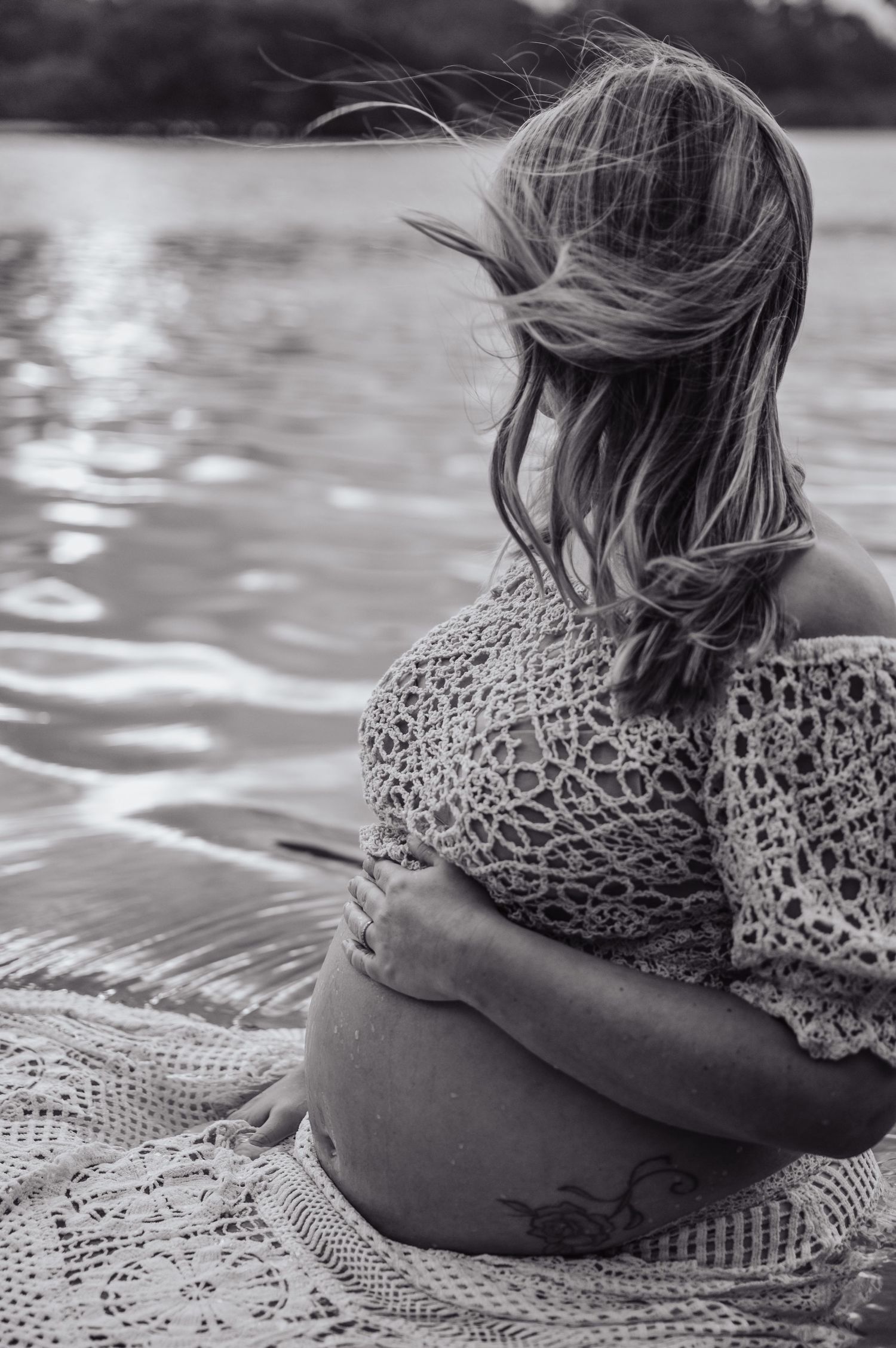 Black and white photo of pregnant woman sitting by water, wind blowing her hair, wearing lace top with tattoo visible.
Maternity Photography