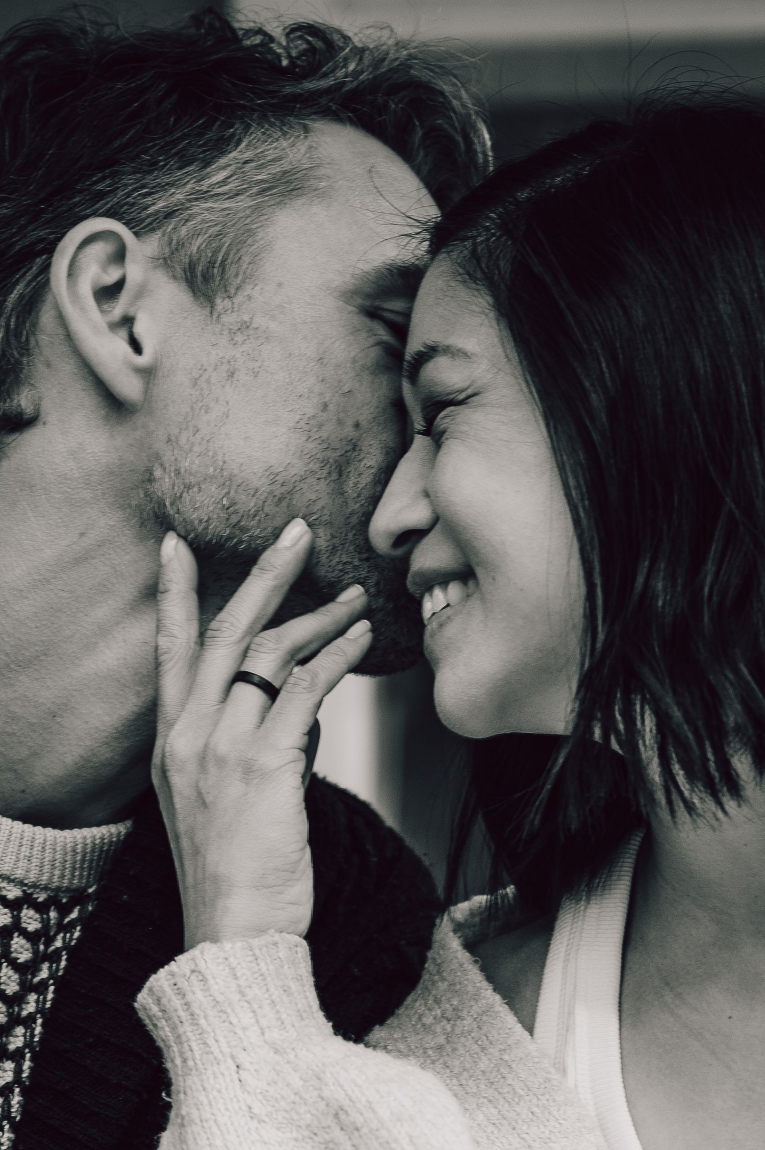 Black and white photo of a smiling couple sharing a tender moment, man kissing womans cheek, wearing a ring. Couples Photography
