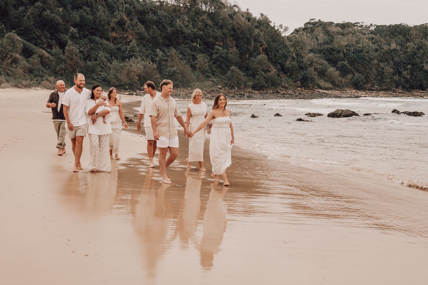 Large family group walking along sandy beach shoreline in coordinating white outfits with lush green hills in background. Family Photography