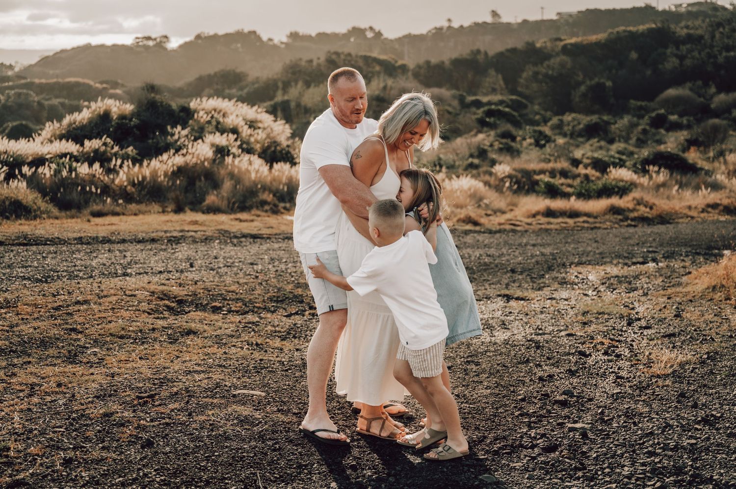 A couple in white outfits embrace with a child on a rocky beach at golden hour with lush greenery behind. Family Photography